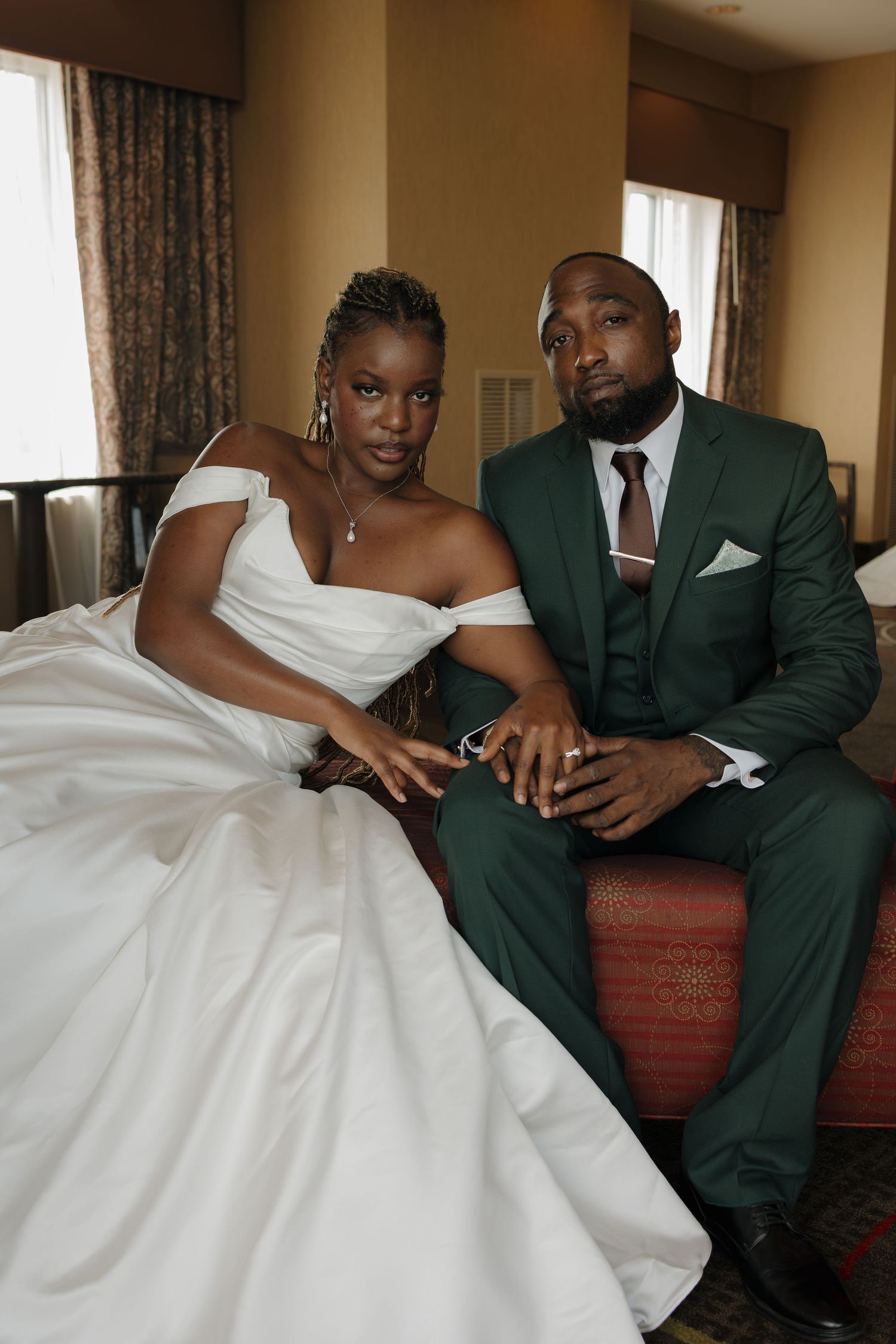 A bride in a white off-shoulder gown and groom in a green suit sit together on a sofa in an elegant room.
