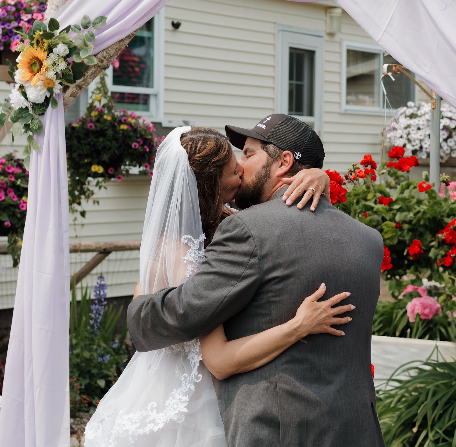 A tender embrace between newlyweds at an outdoor wedding ceremony with white drapes and colorful flowers in the background.