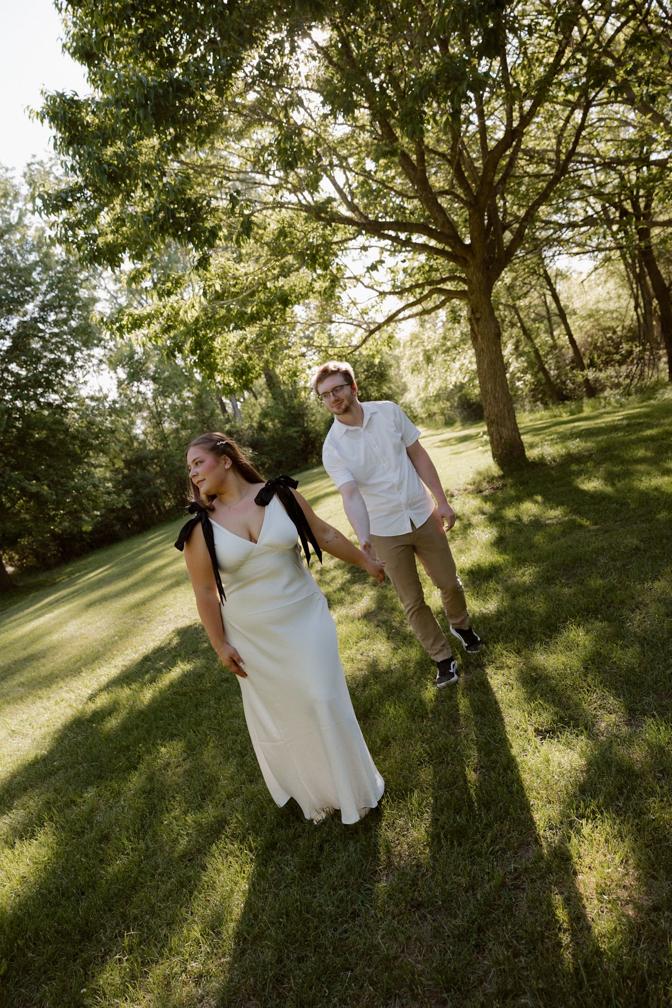 A bride in a white gown and groom in casual attire walk hand-in-hand through a lush green park on their wedding day.