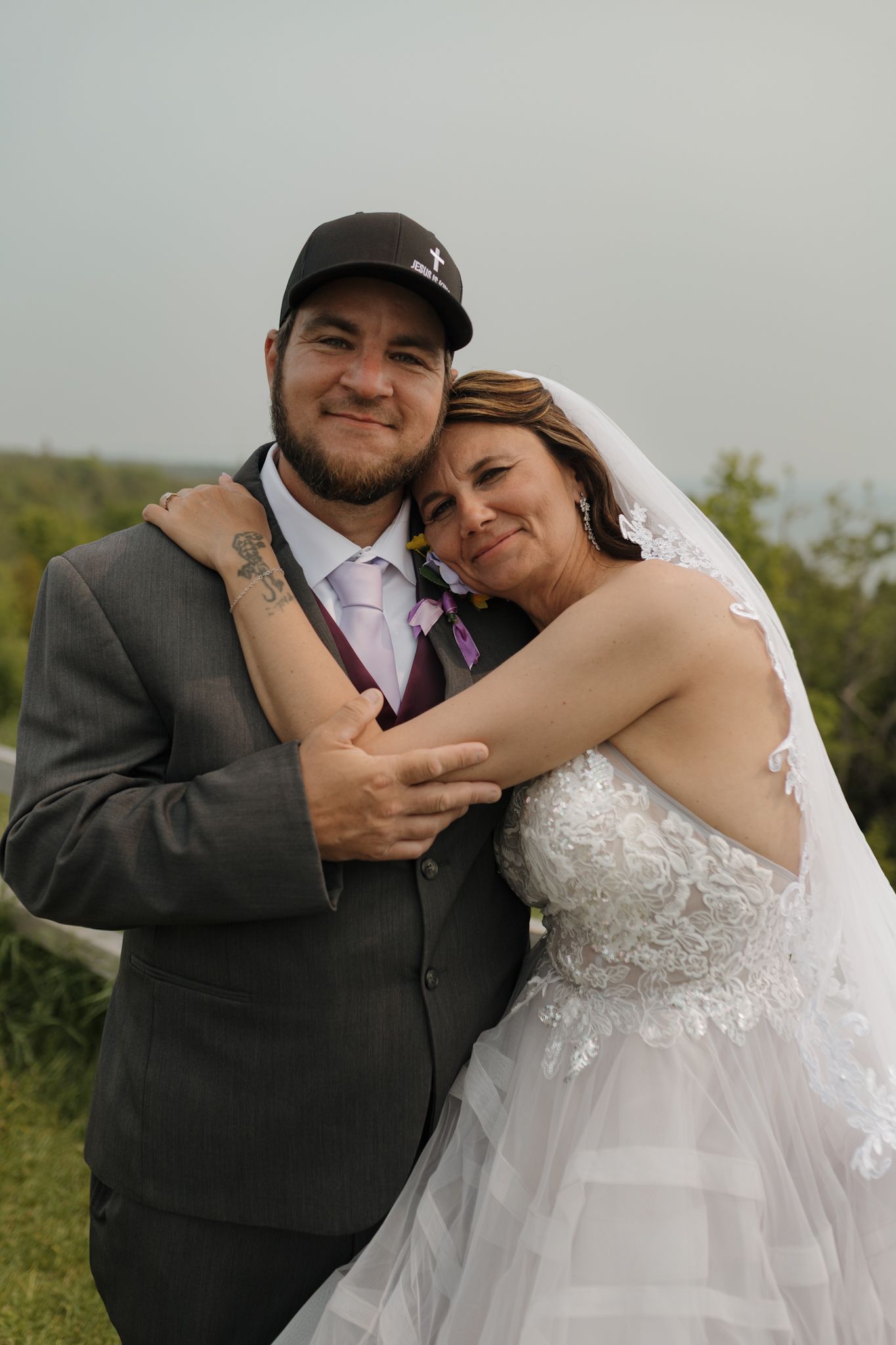Happy newlyweds embrace outdoors, bride in lace gown with veil, groom in gray suit and cap, sharing a joyful wedding moment.