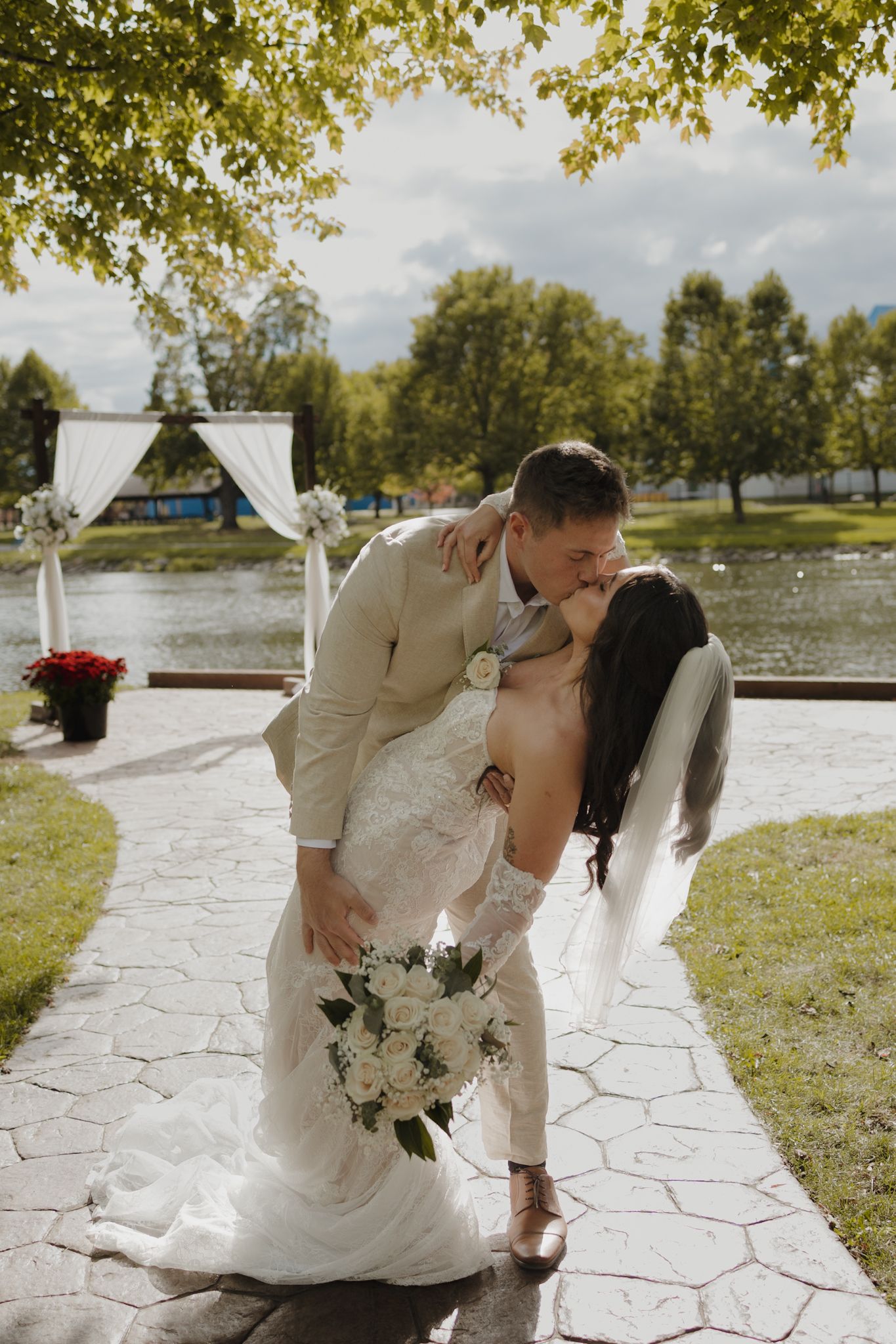 A romantic wedding couple shares a kiss on a stone patio with a lakeside view on a sunny day.