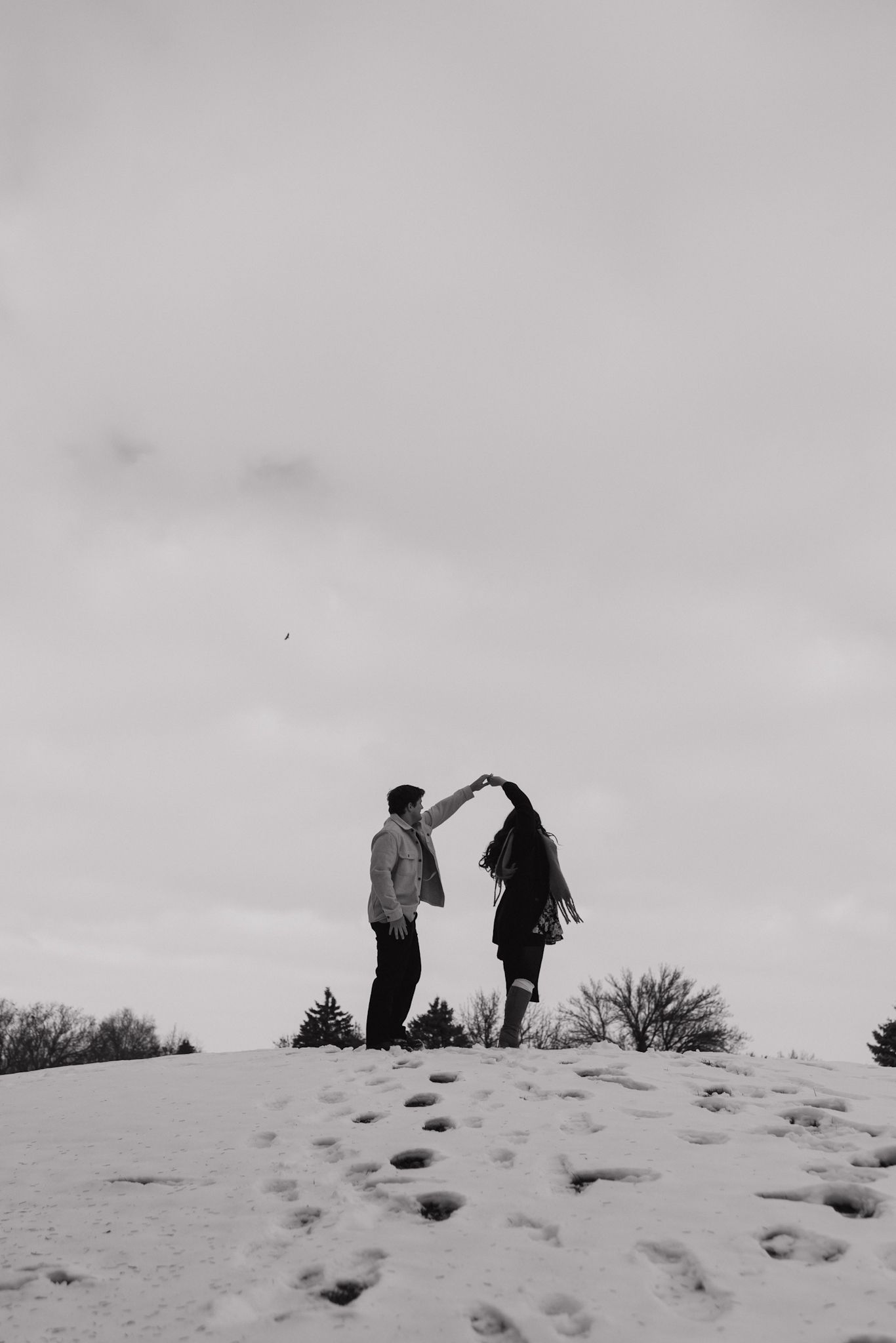 Two silhouettes stand atop a snowy hill against an overcast sky, forming heart shapes with their hands.