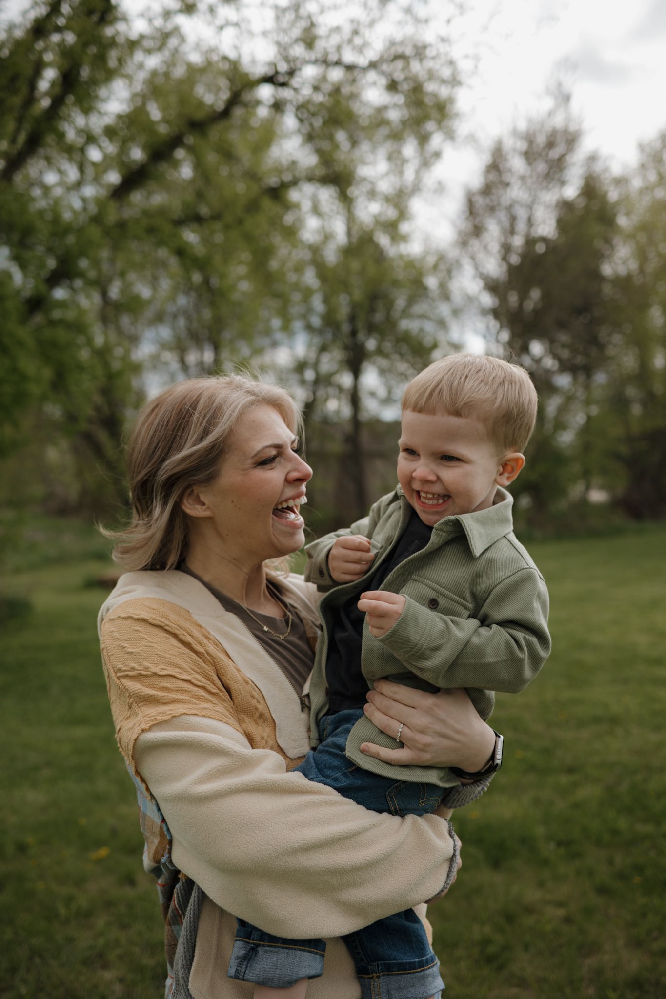 A smiling mother holds her laughing toddler boy outdoors in a lush green park setting.
