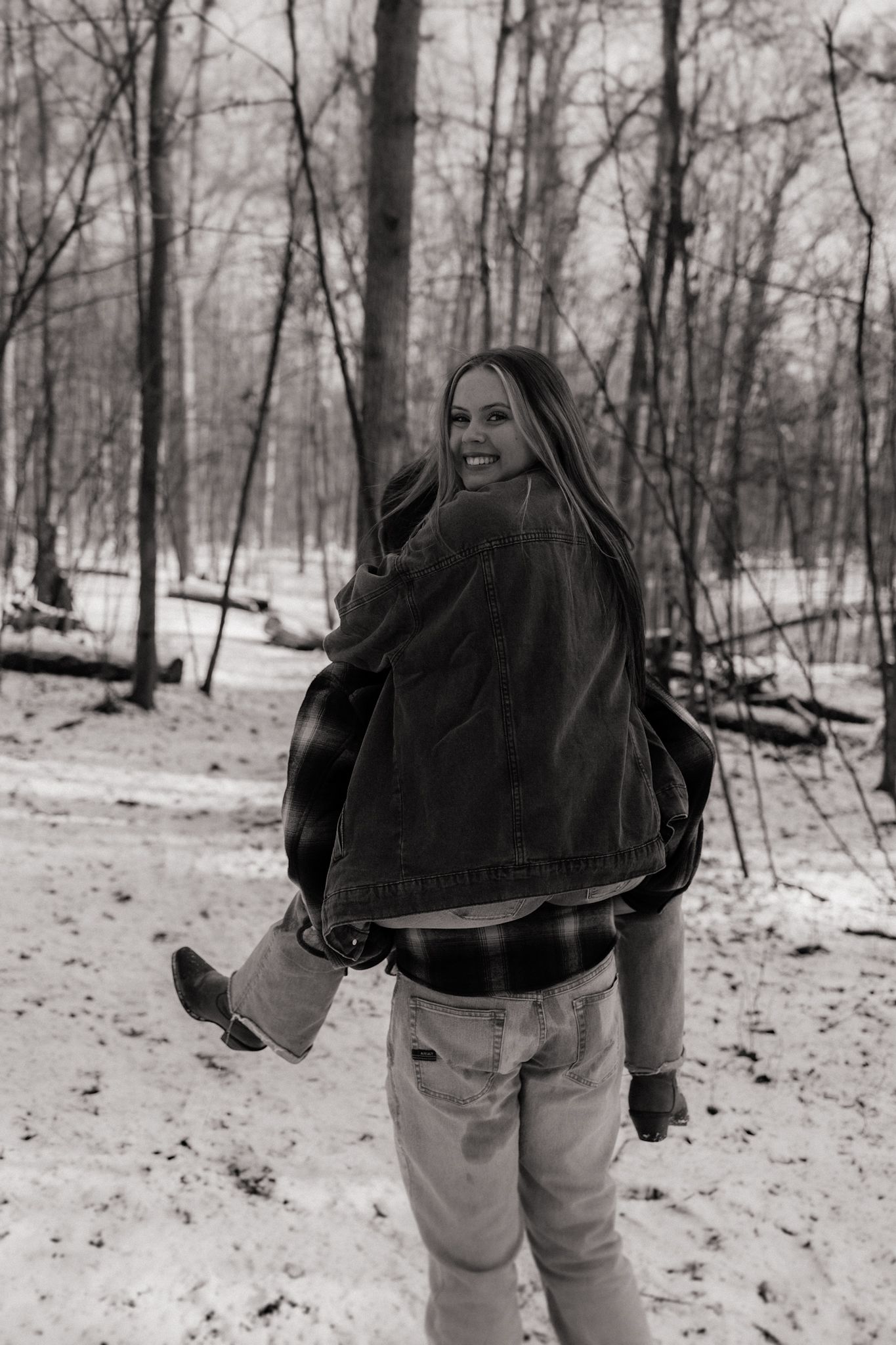 Black and white winter scene of person in denim and jacket looking back while walking through snowy forest.