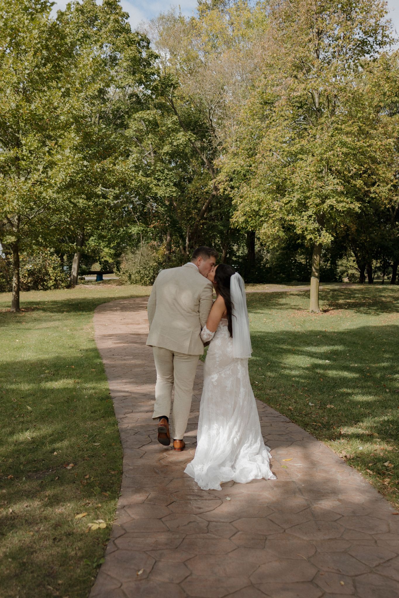A couple in wedding attire stands together on a stone path surrounded by lush green trees in a park setting.