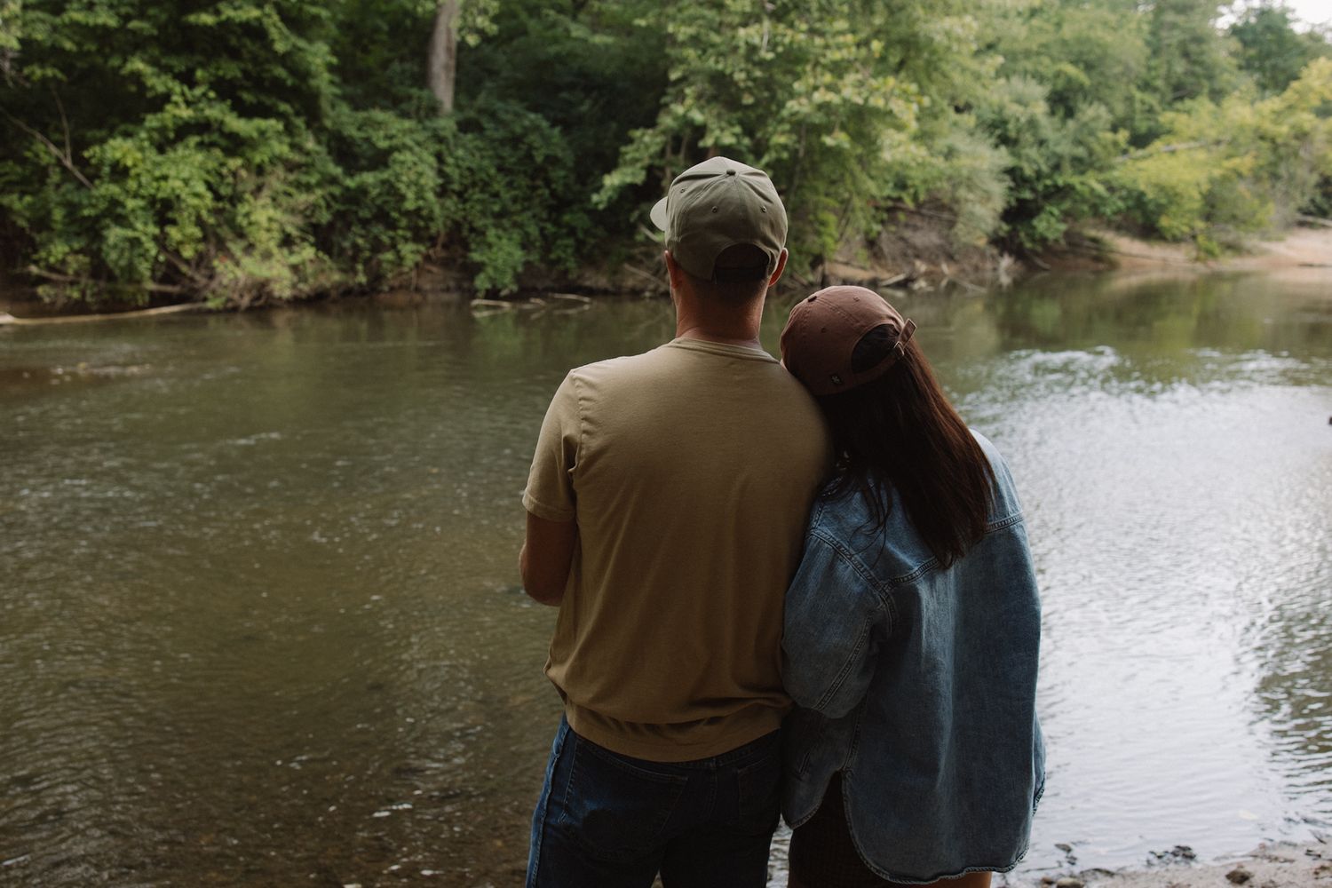 Couple standing together by a peaceful river surrounded by lush green trees, viewed from behind.