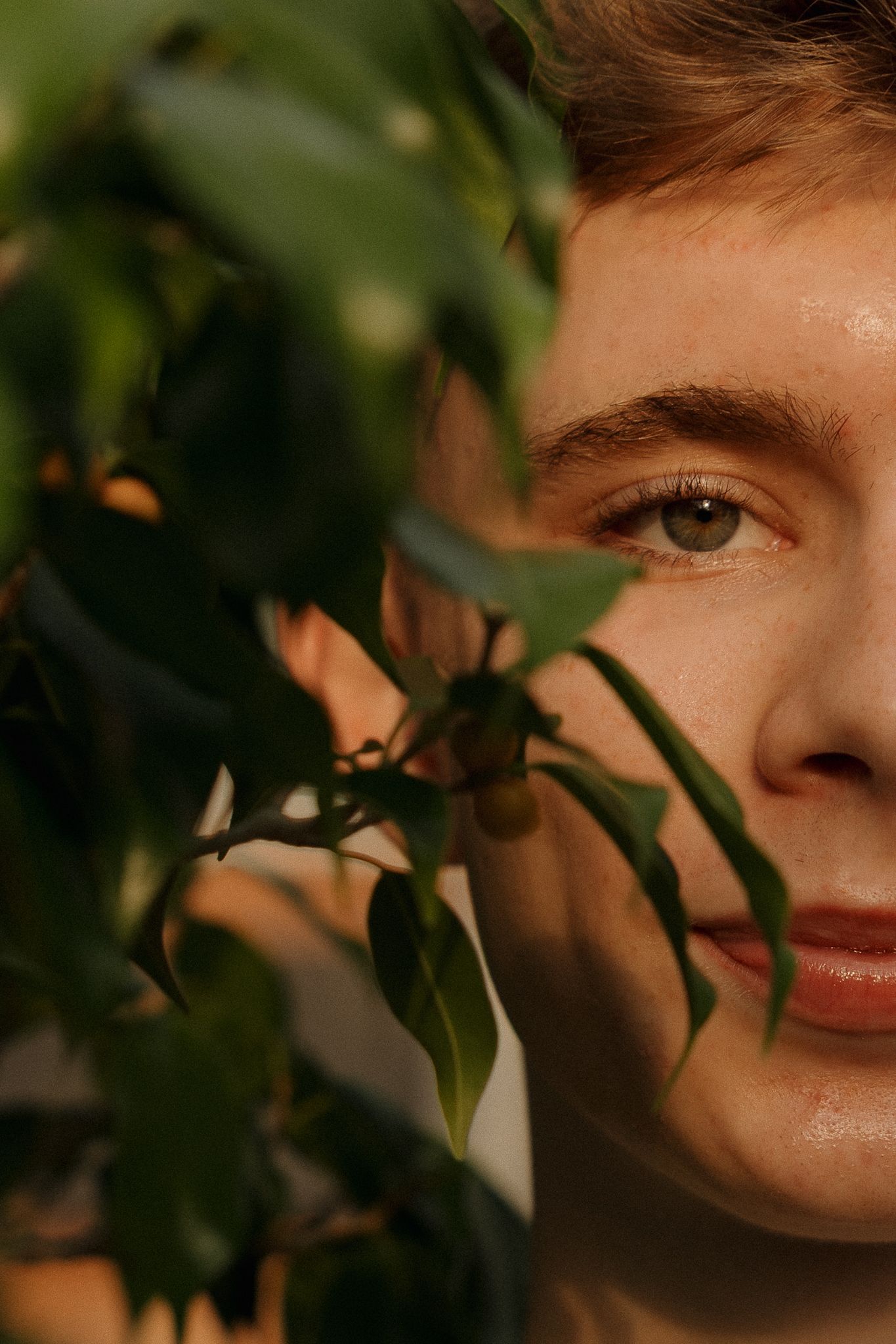 Close-up portrait of a young man partially hidden behind green plant leaves in warm natural light.