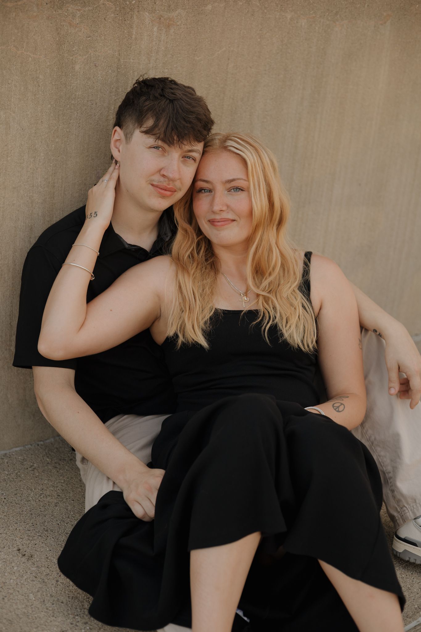 A couple in black outfits poses intimately for a romantic photoshoot against a neutral wall.