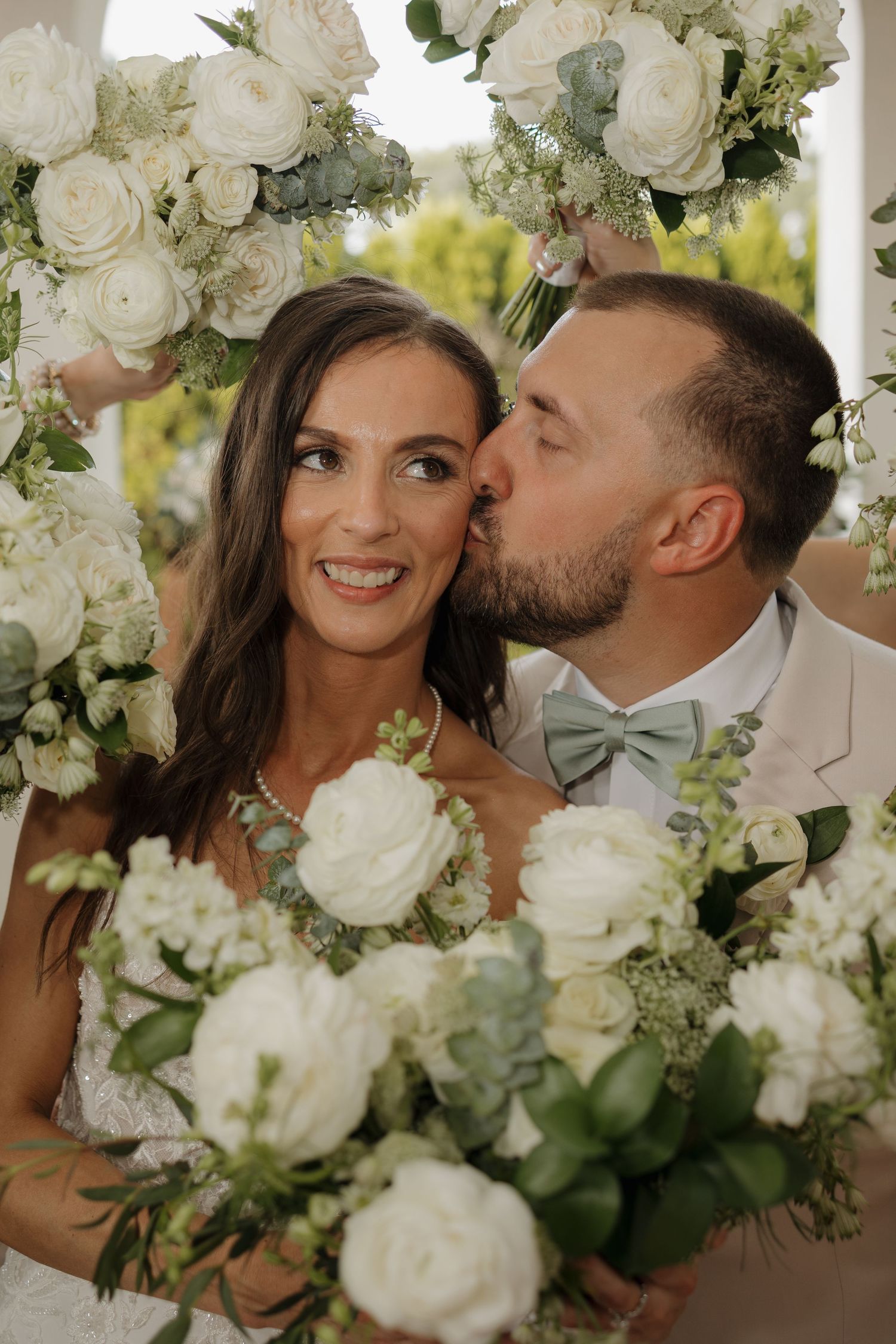 Bride smiling as groom kisses her cheek, surrounded by white rose bouquets and floral arch at wedding ceremony.