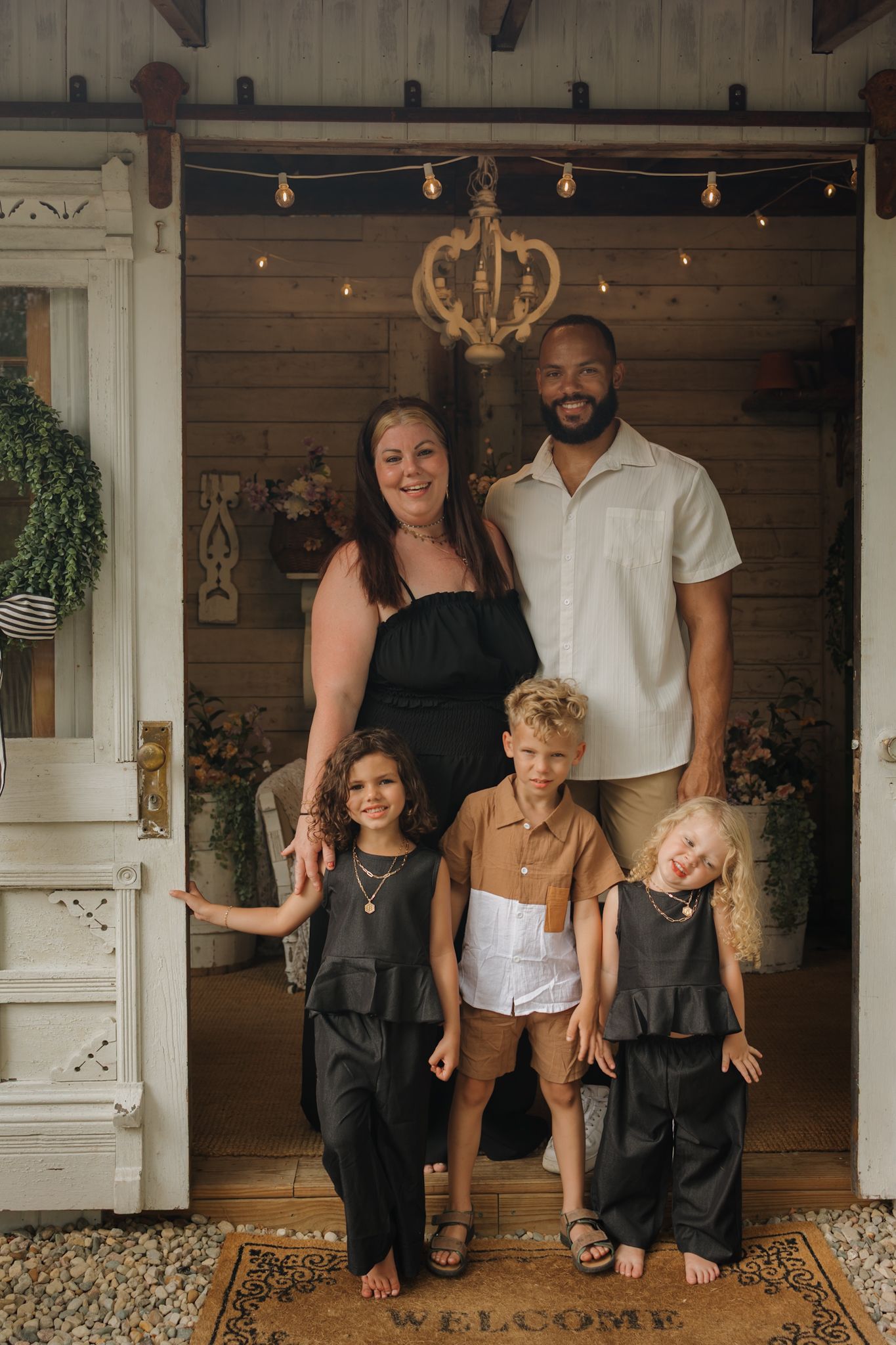 Family of five posing on a welcoming porch with string lights, parents standing behind three young children on a welcome mat.