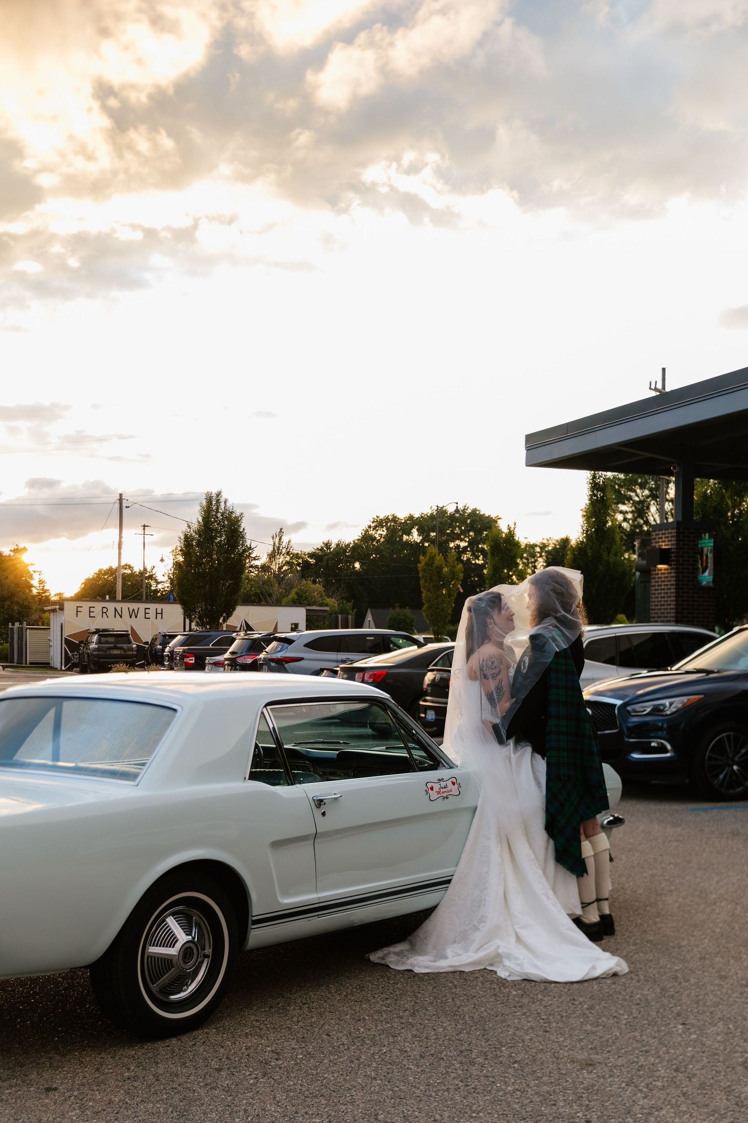 A classic white convertible car serves as a romantic backdrop for a wedding couple at sunset.