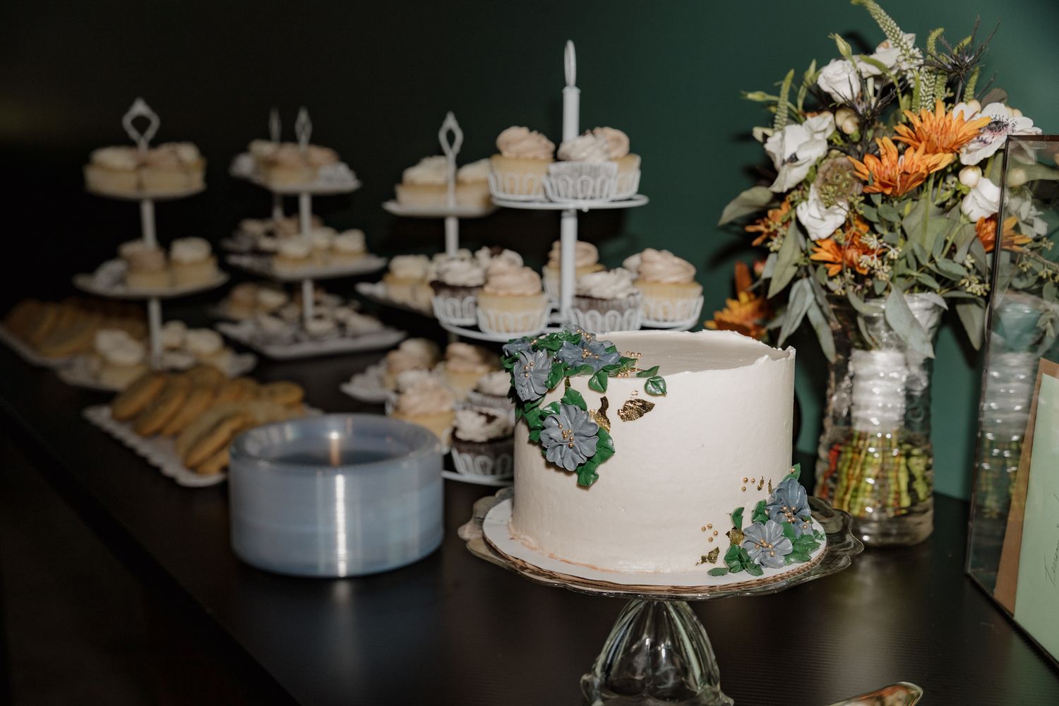 Elegant wedding dessert table with tiered white cakes, floral decorations, and vintage-style cake stands on dark backdrop.