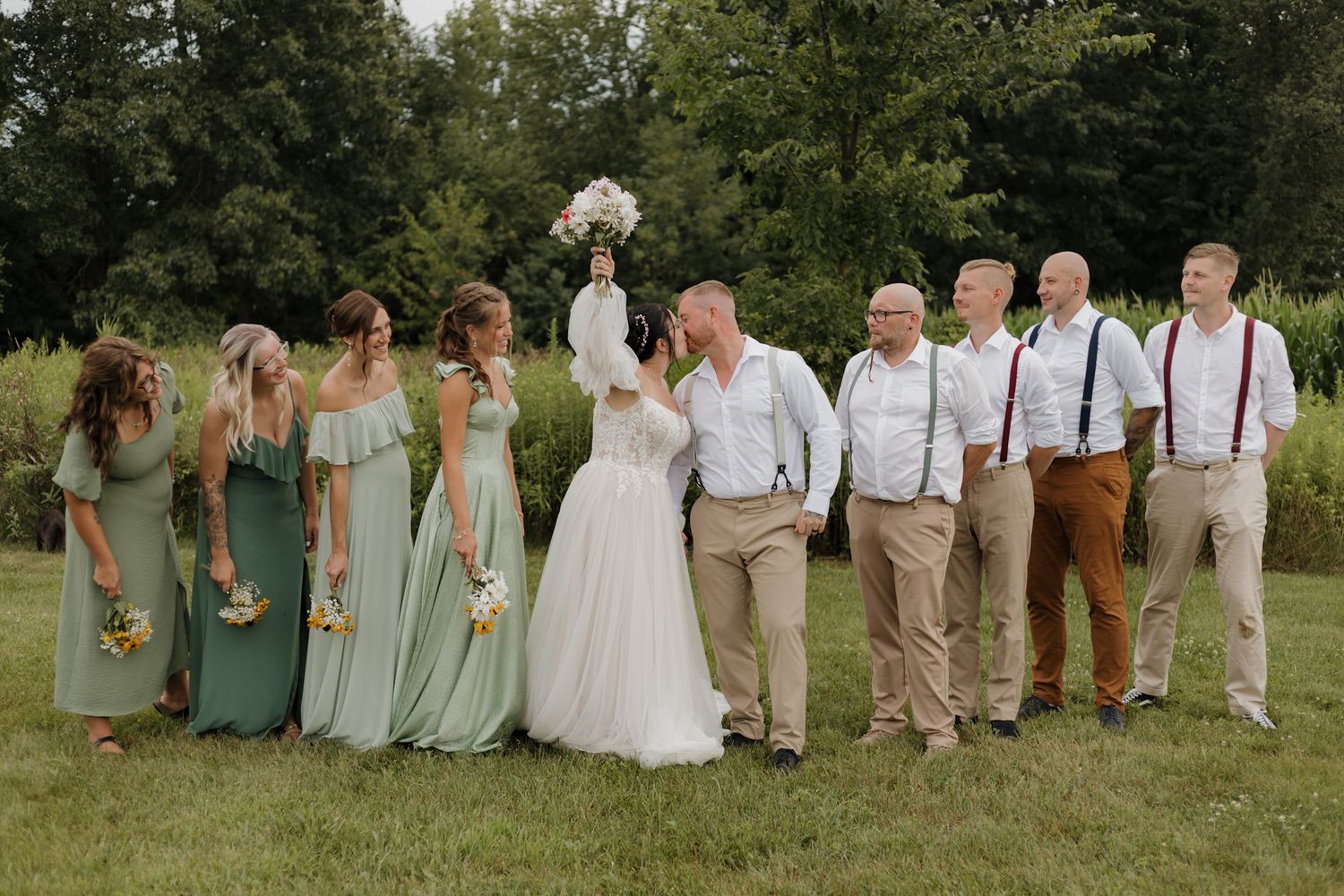 Wedding party in sage green dresses and khaki suits poses together on grassy lawn during summer celebration.