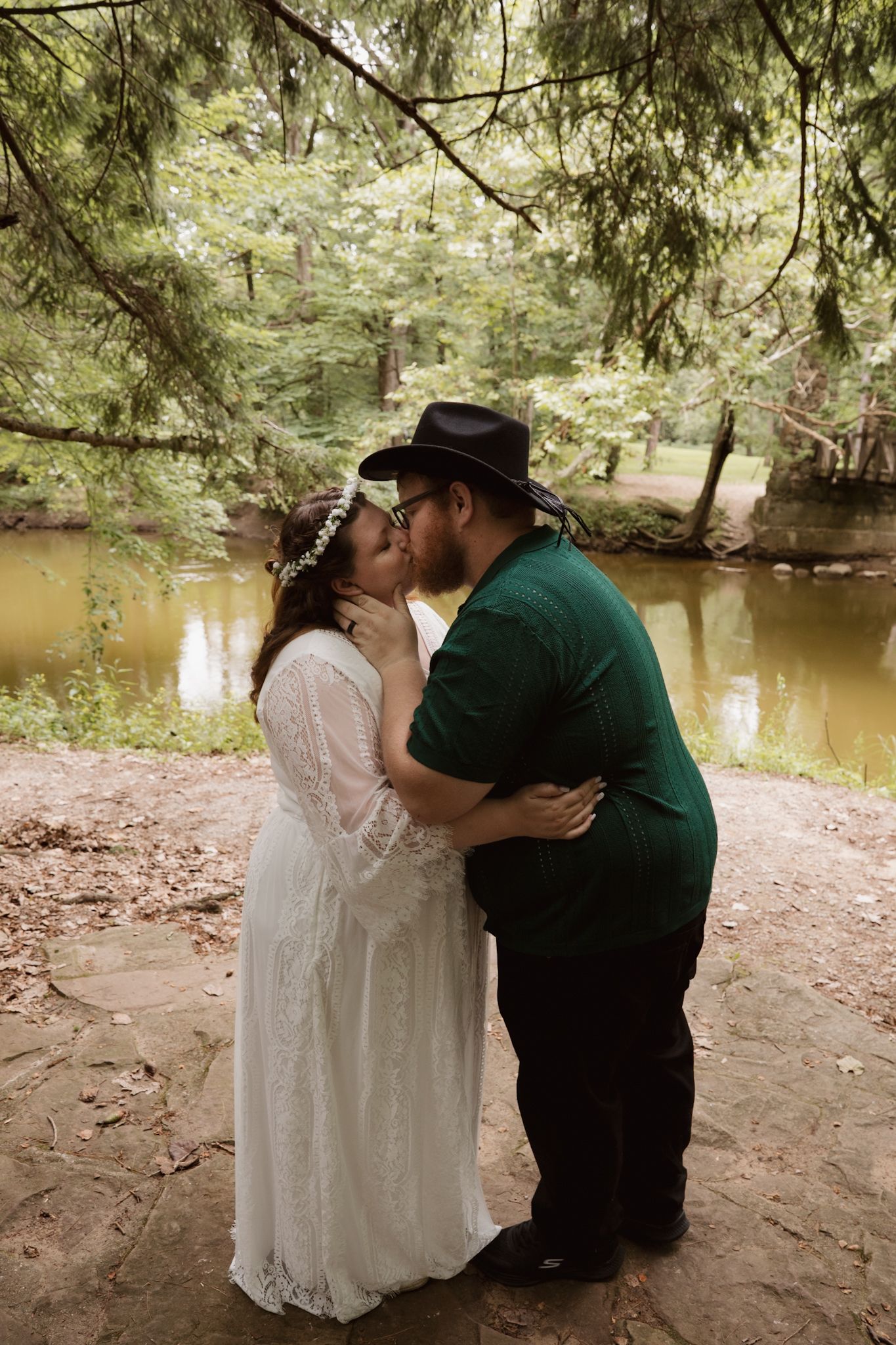 A couple shares a tender moment by a serene riverbank surrounded by overhanging trees.