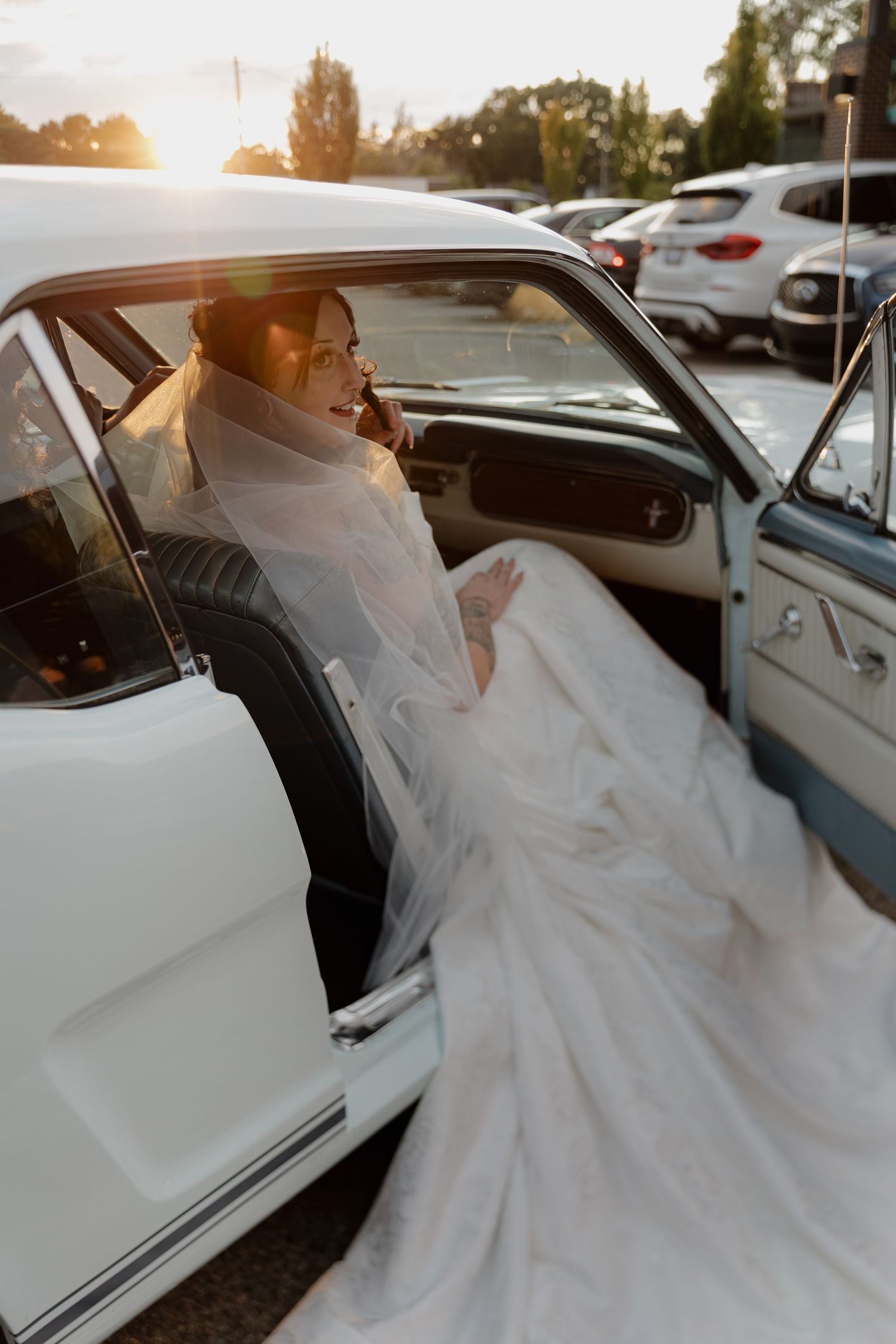 A bride in a flowing white wedding gown gracefully enters a classic white car at sunset.