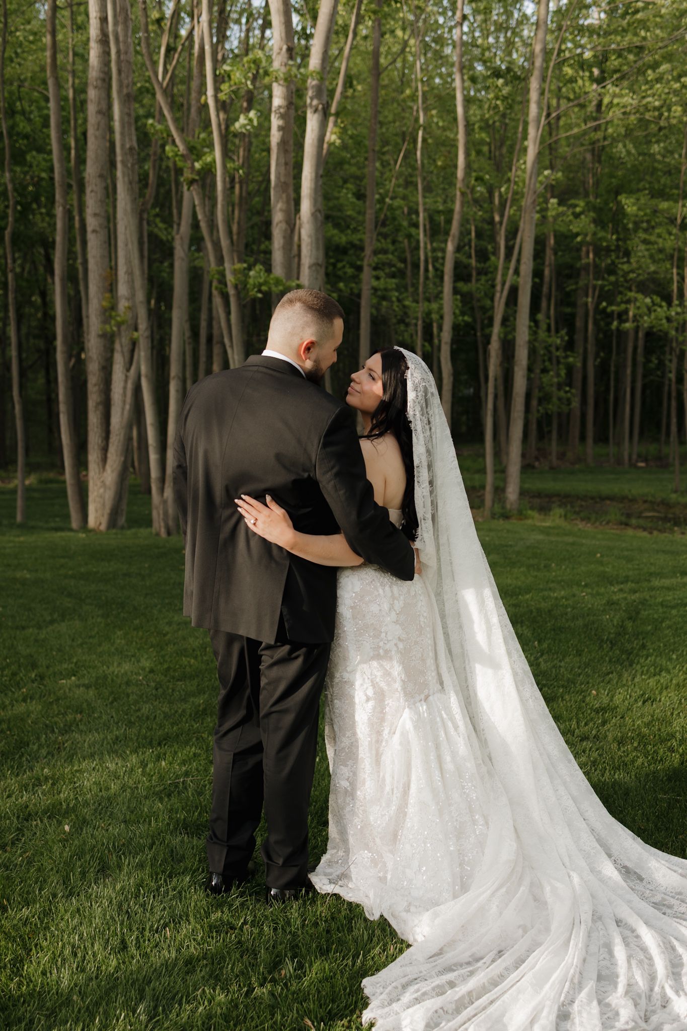 A bride and groom embrace outdoors on green grass with tall trees in the background at their wedding.