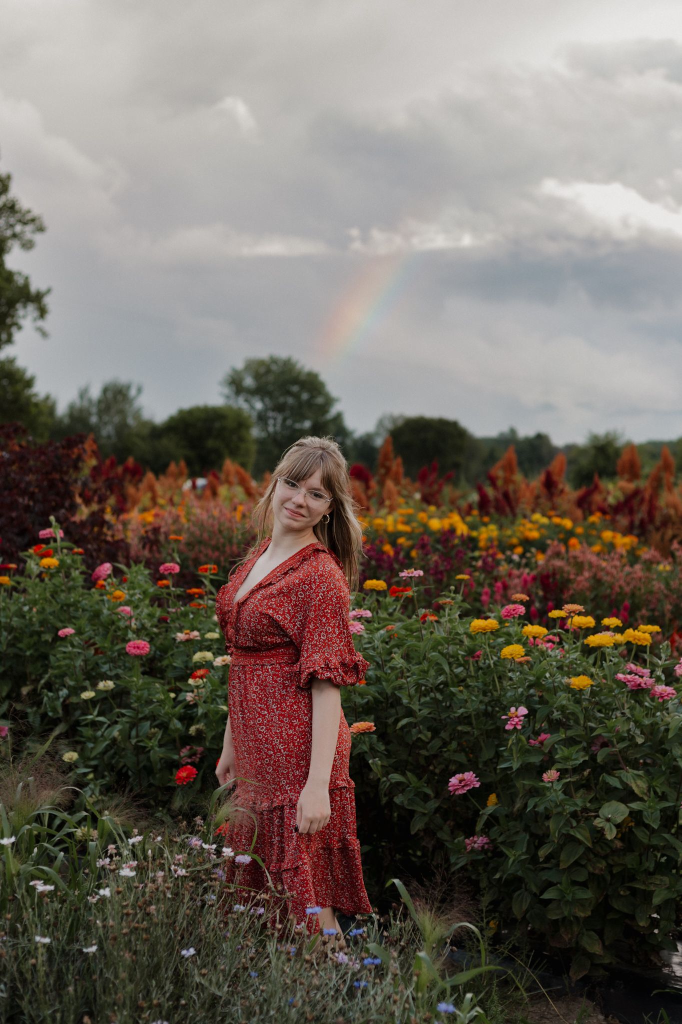 Person in red floral dress walking through colorful flower garden under cloudy sky.