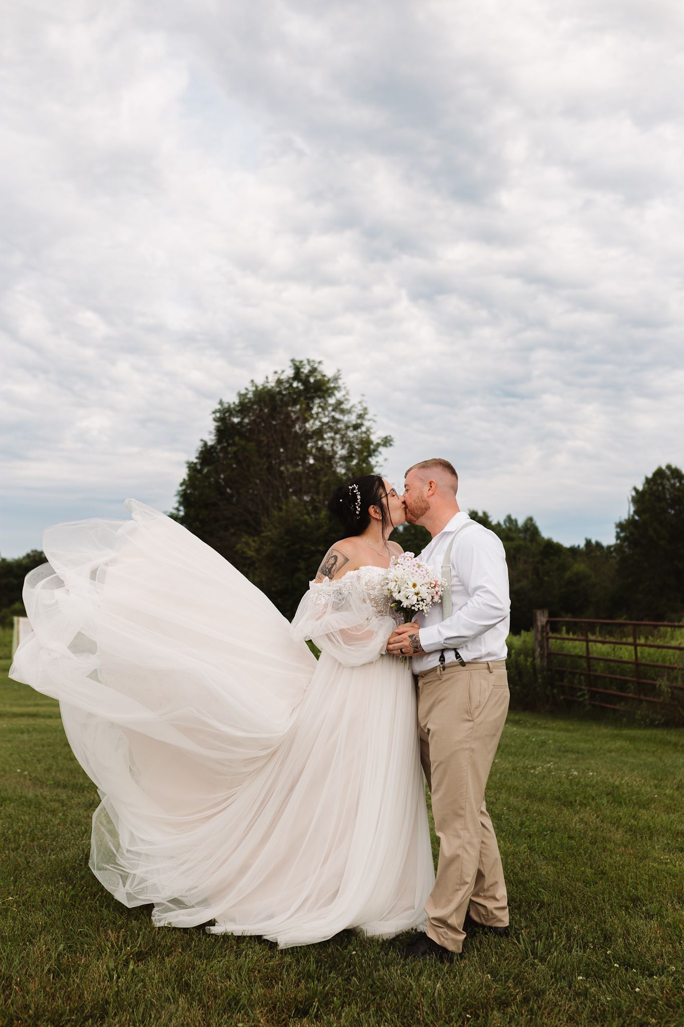 Bride and groom kissing outdoors on green grass, white wedding dress veil flowing in wind, rustic fence and trees background.