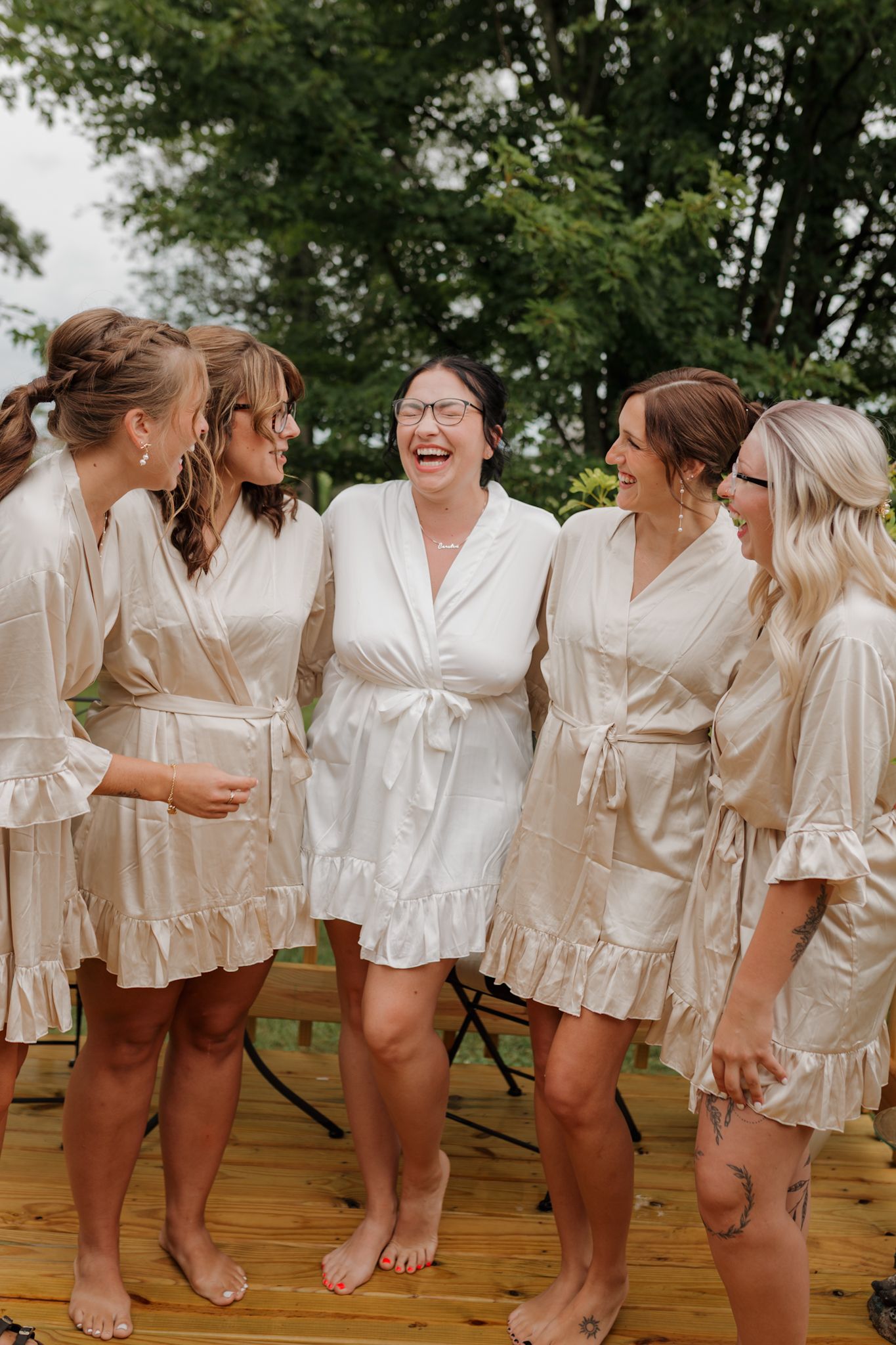 A group of women in matching cream-colored satin robes laugh together while standing barefoot on a wooden deck outdoors.