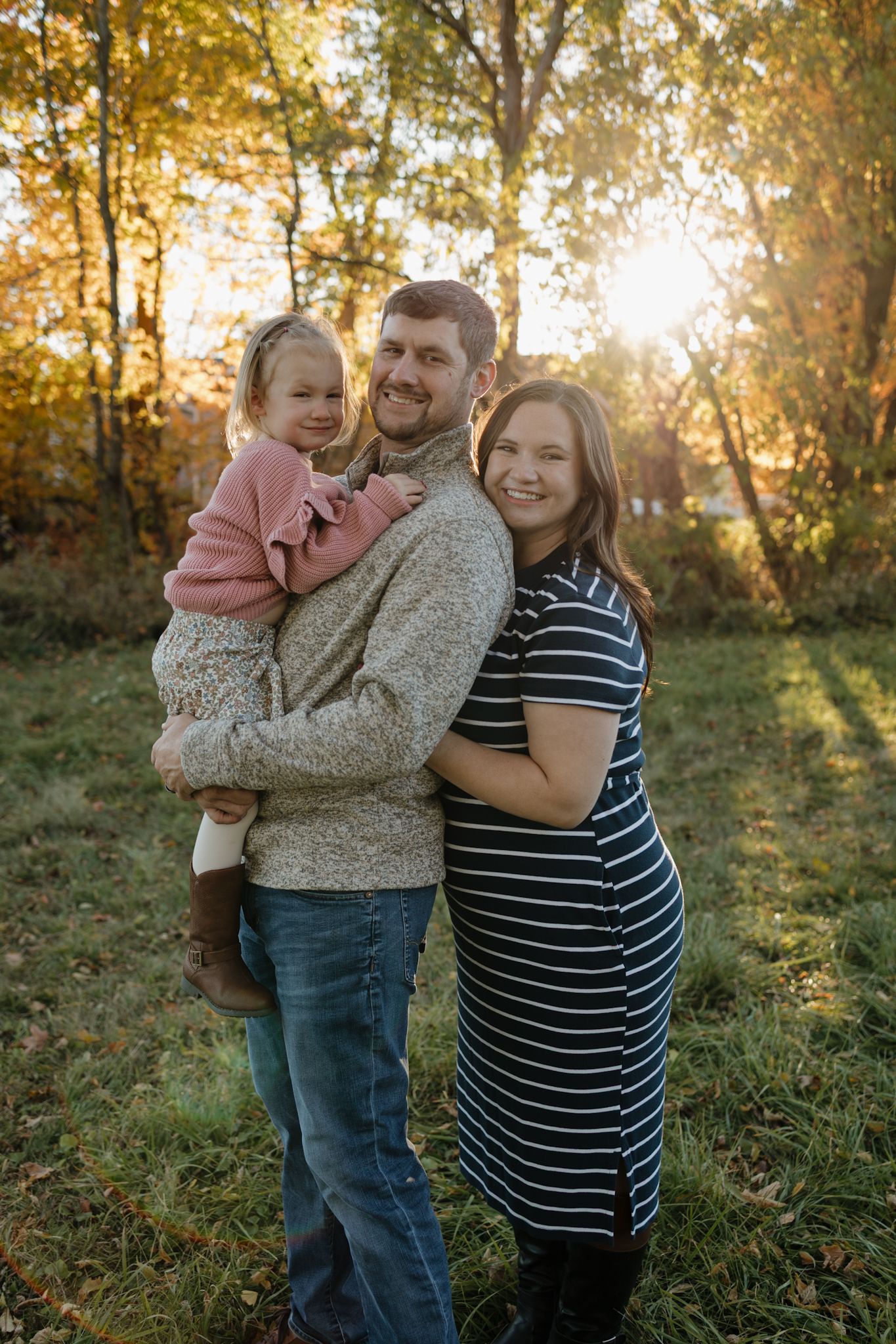 Family of three posing outdoors in autumn, man holding toddler girl, woman in striped dress, golden fall foliage background.