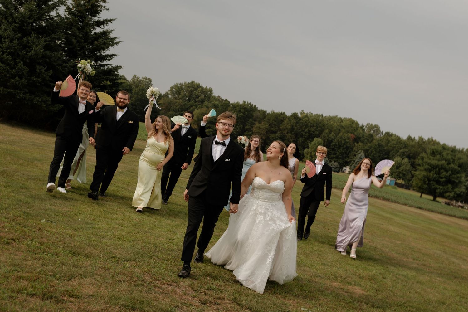 Bride and groom walk hand-in-hand with cheering wedding party on green lawn at outdoor wedding celebration.