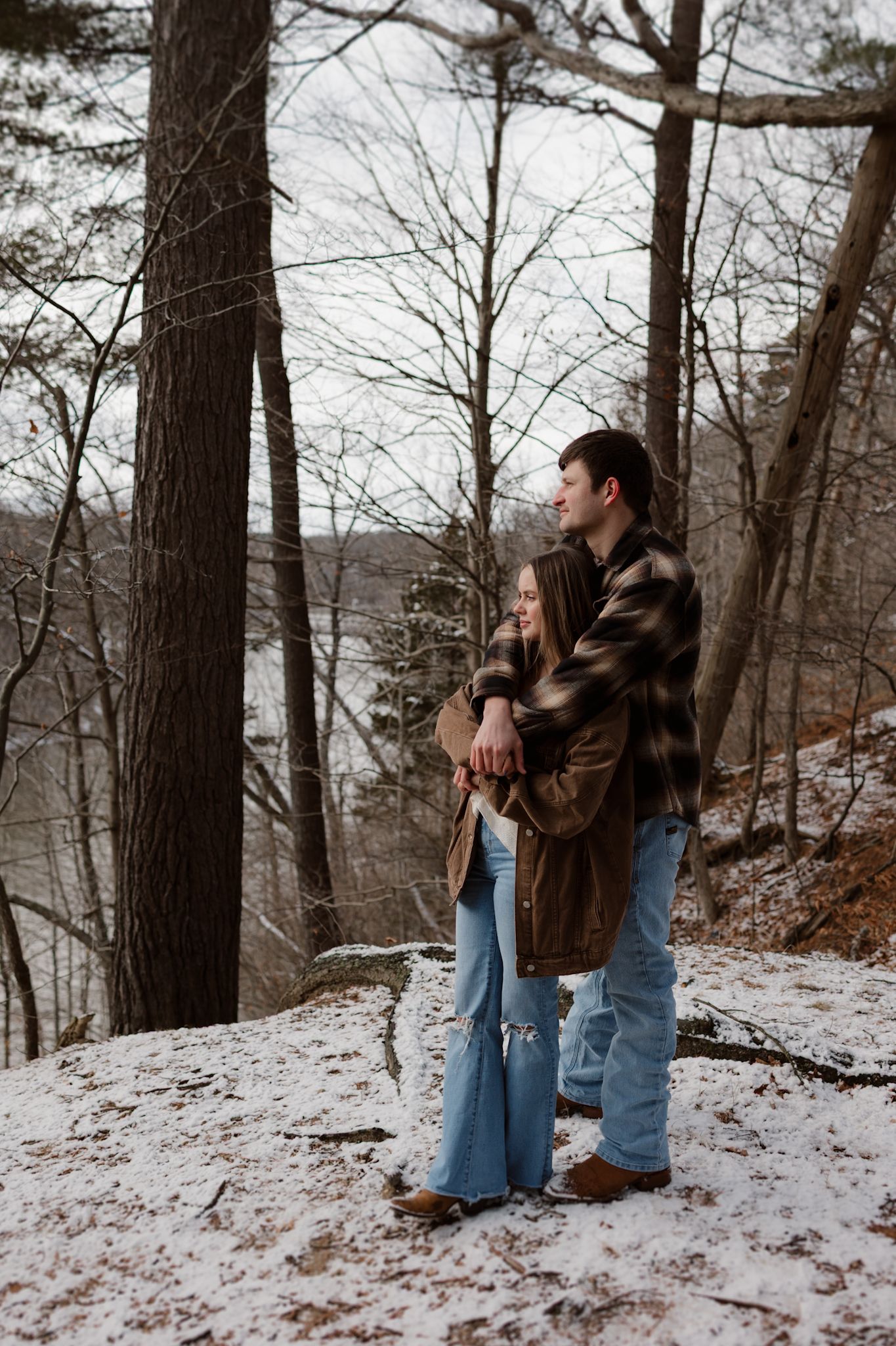 A couple embraces in a snowy winter forest setting wearing casual clothing and brown jackets.