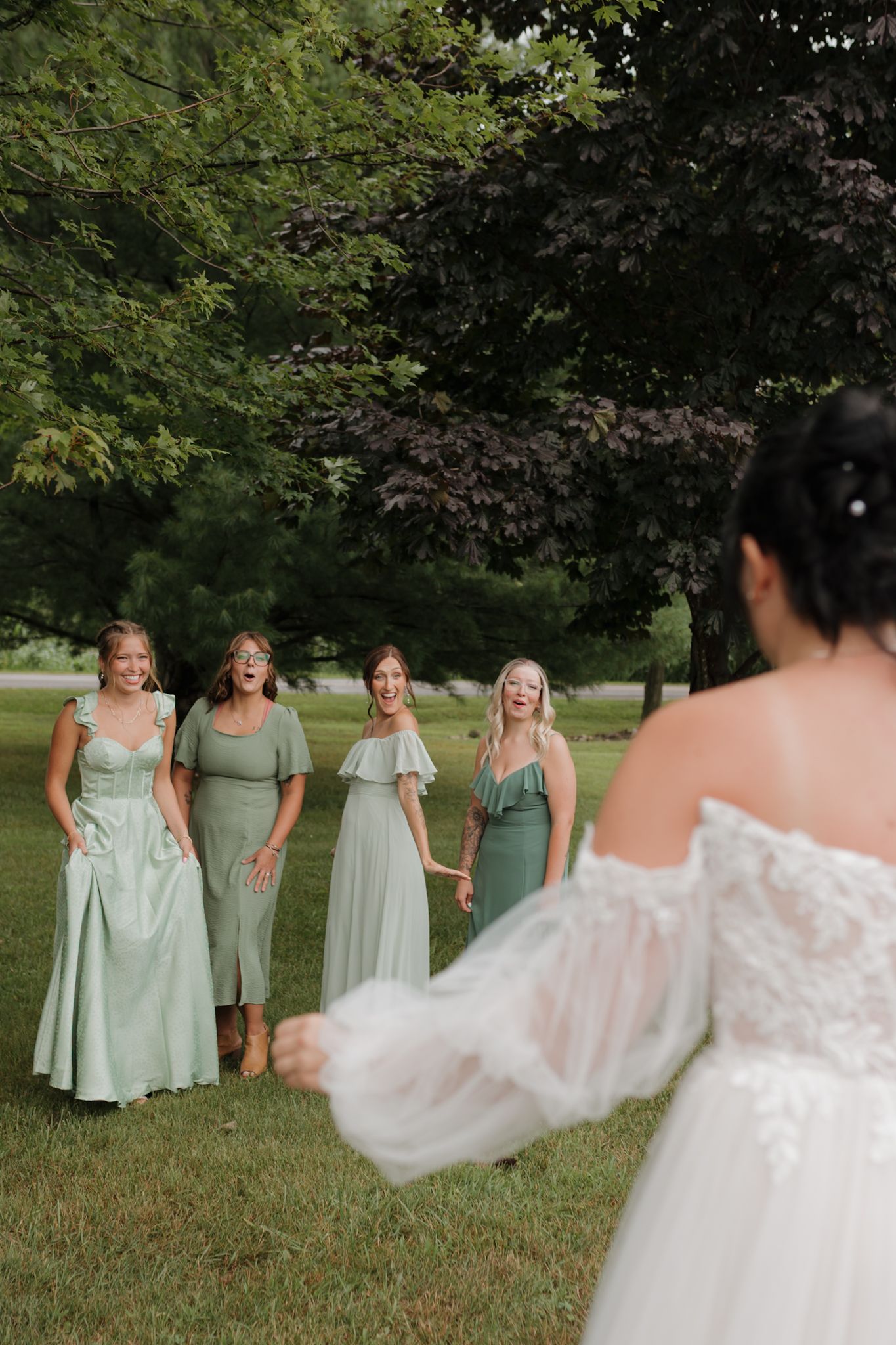 Bridesmaids in sage green dresses share a candid moment outdoors with the bride in a flowing white gown.