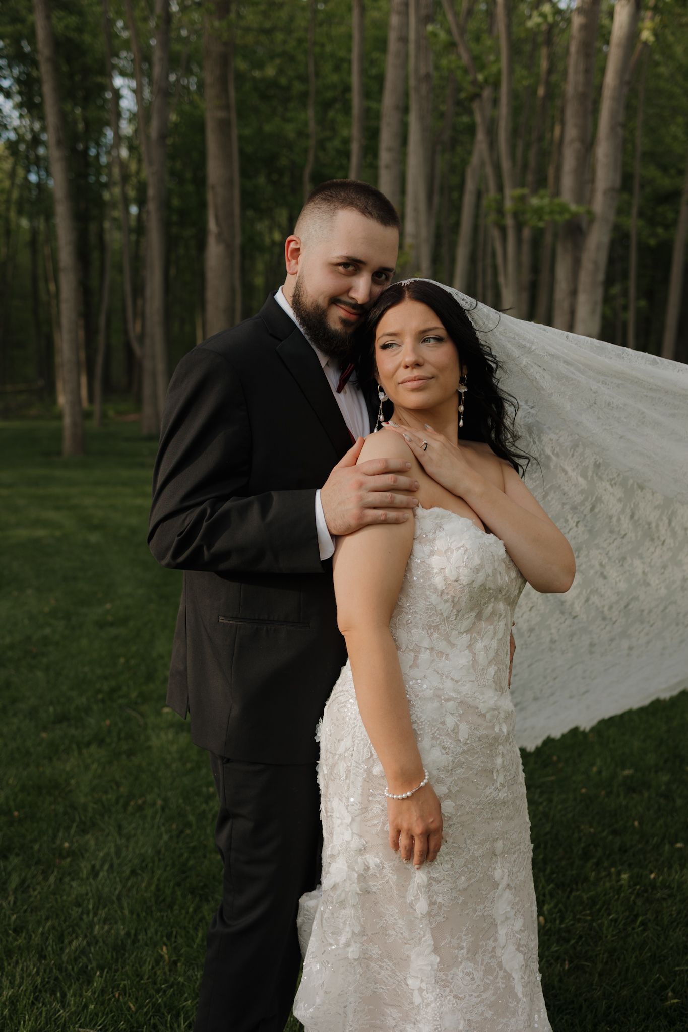 Bride and groom embrace outdoors among tall trees, her lace gown and flowing veil catching the breeze on their wedding day.