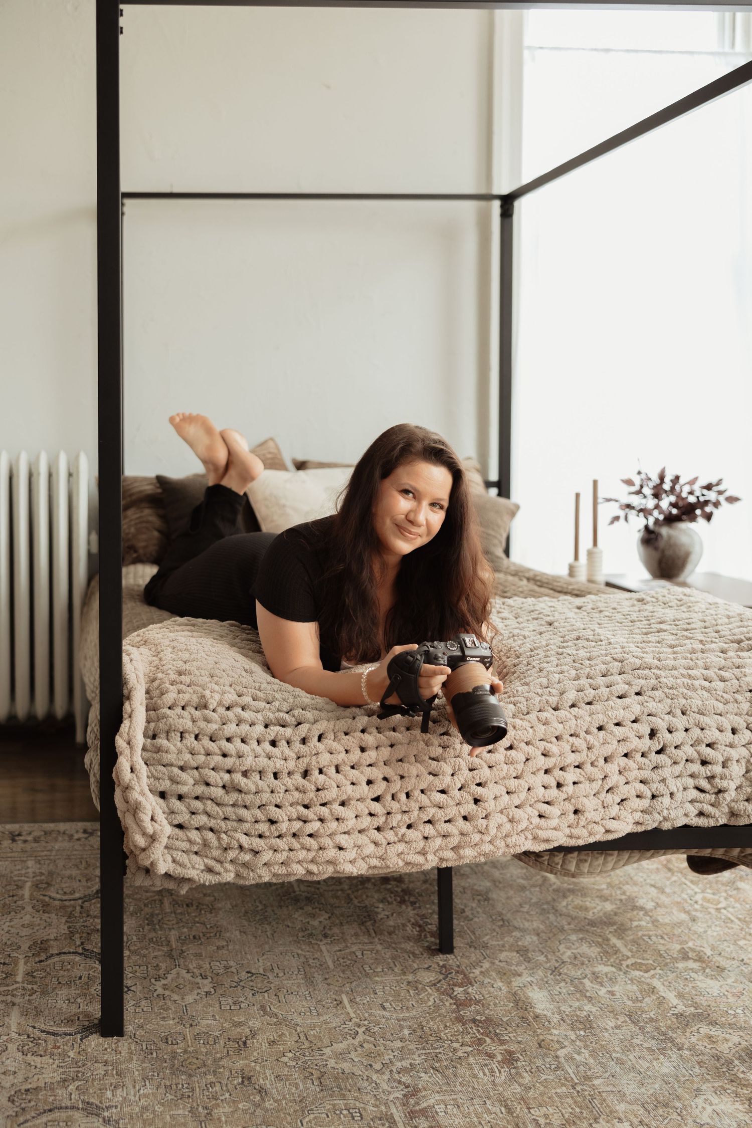 Woman in black outfit lying on a four-poster bed holding a camera, with chunky knit blanket in a bright bedroom.