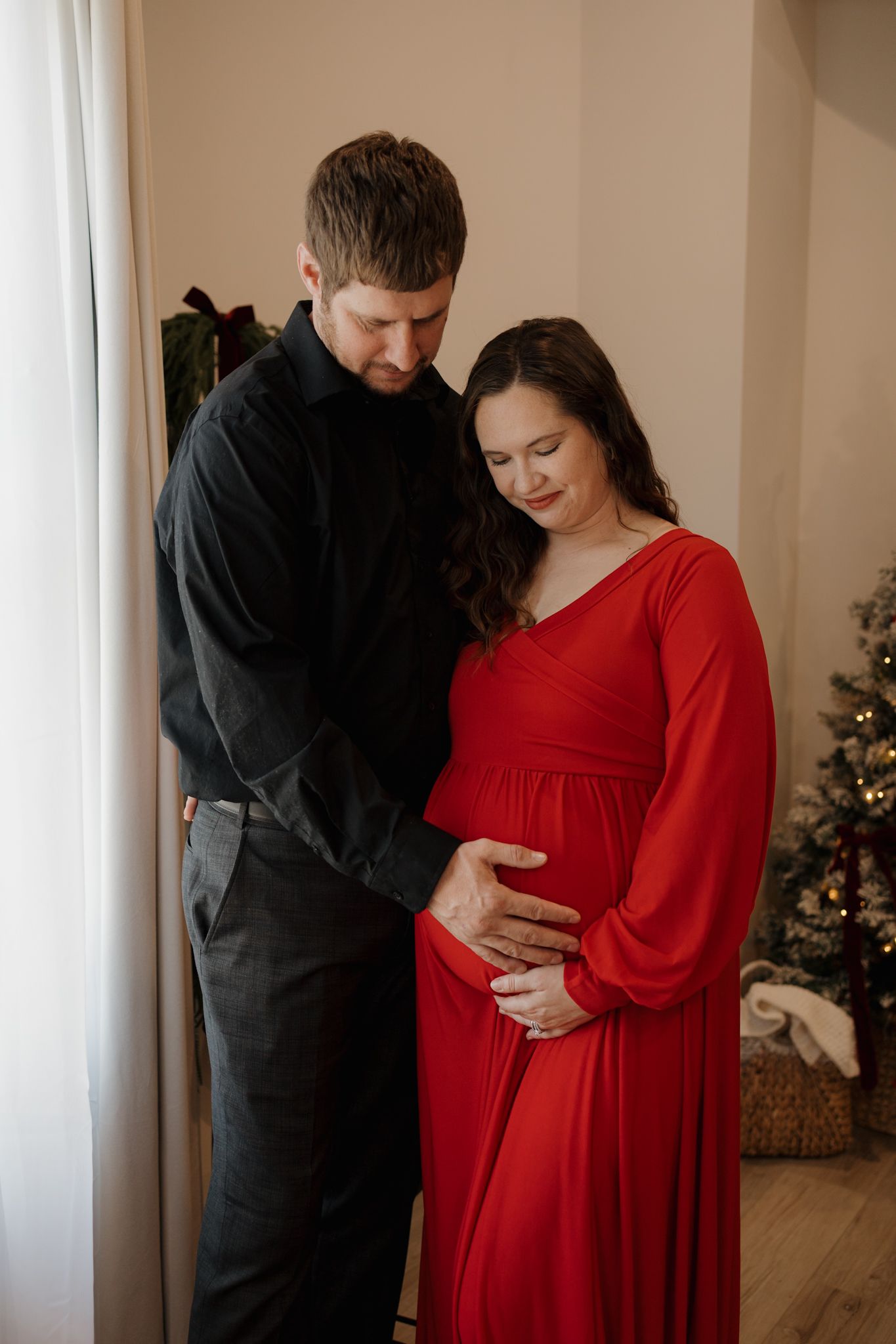 A couple in black and red attire share intimate moments during a maternity photoshoot near a Christmas tree.