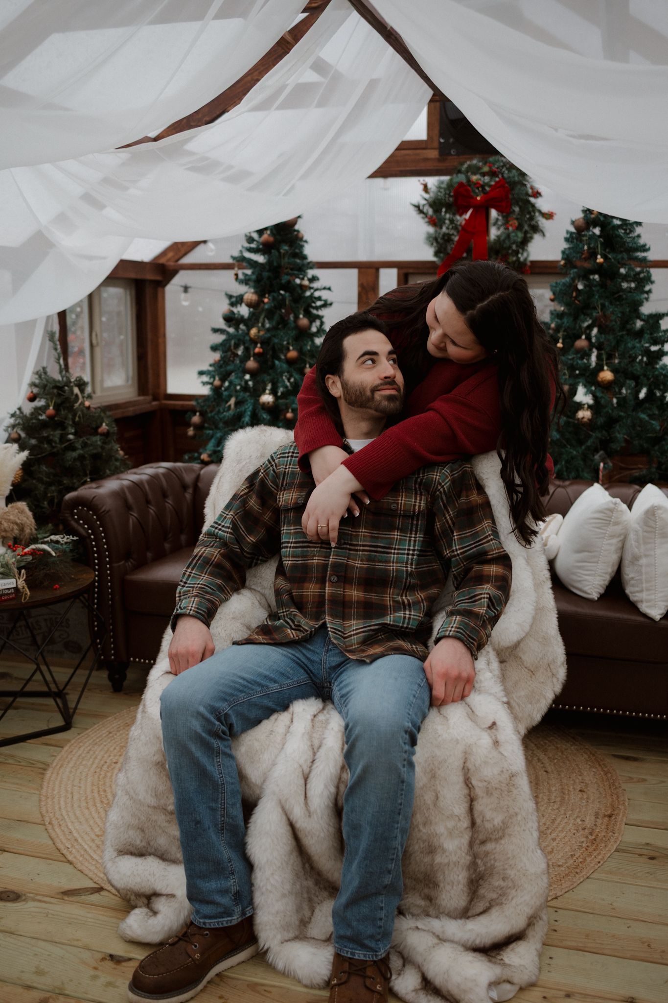 A couple sits on a couch with their white fluffy dog during a cozy Christmas photoshoot with decorated trees in background.