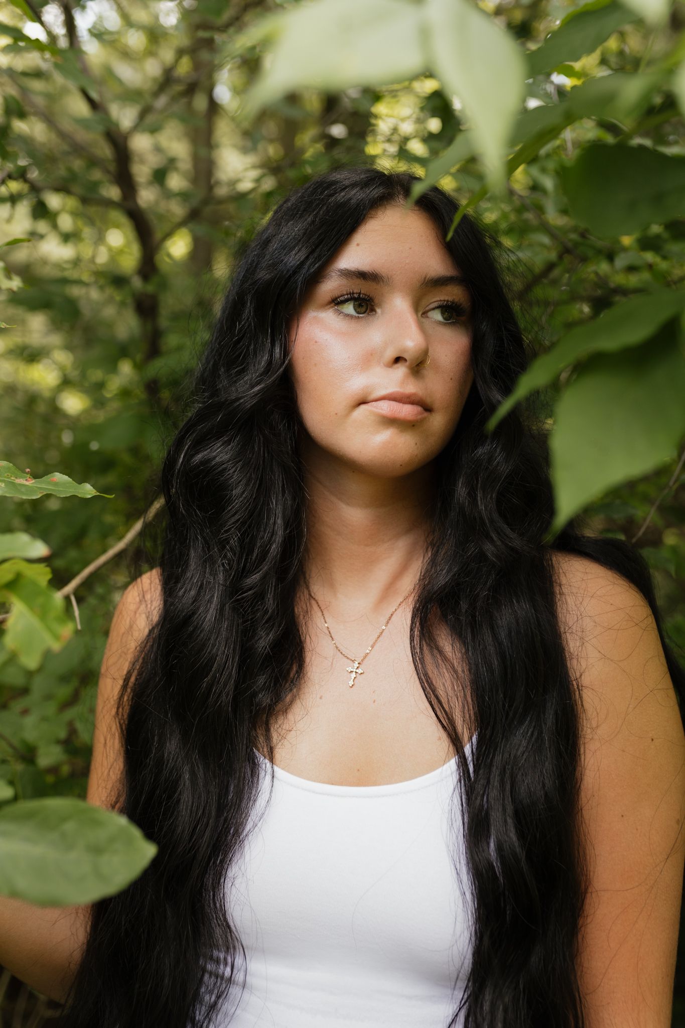 Portrait in a white tank top surrounded by green foliage on a sunny day.