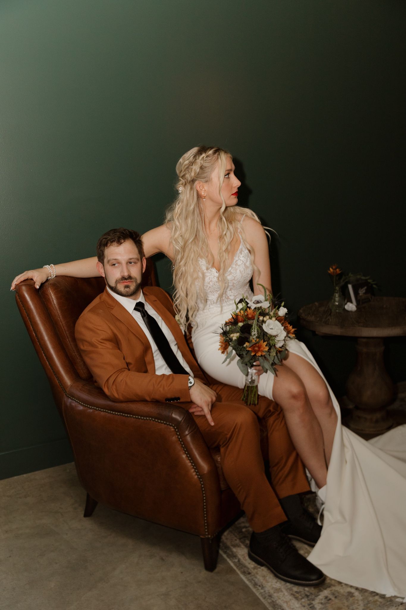 A couple in wedding attire poses intimately on a brown leather chair against a dark green backdrop.