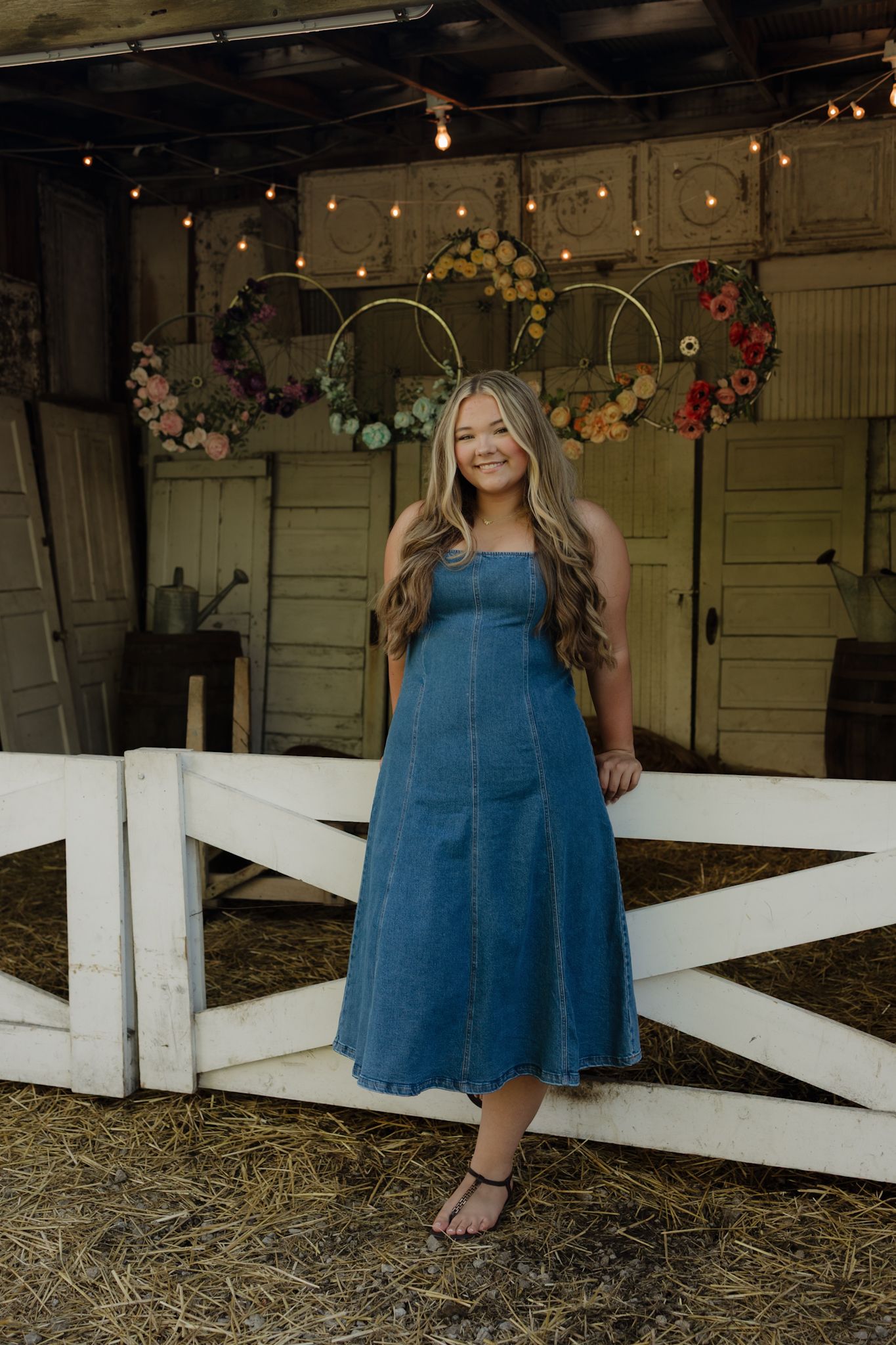Young woman in denim midi dress smiling in rustic barn with floral wreaths and string lights.