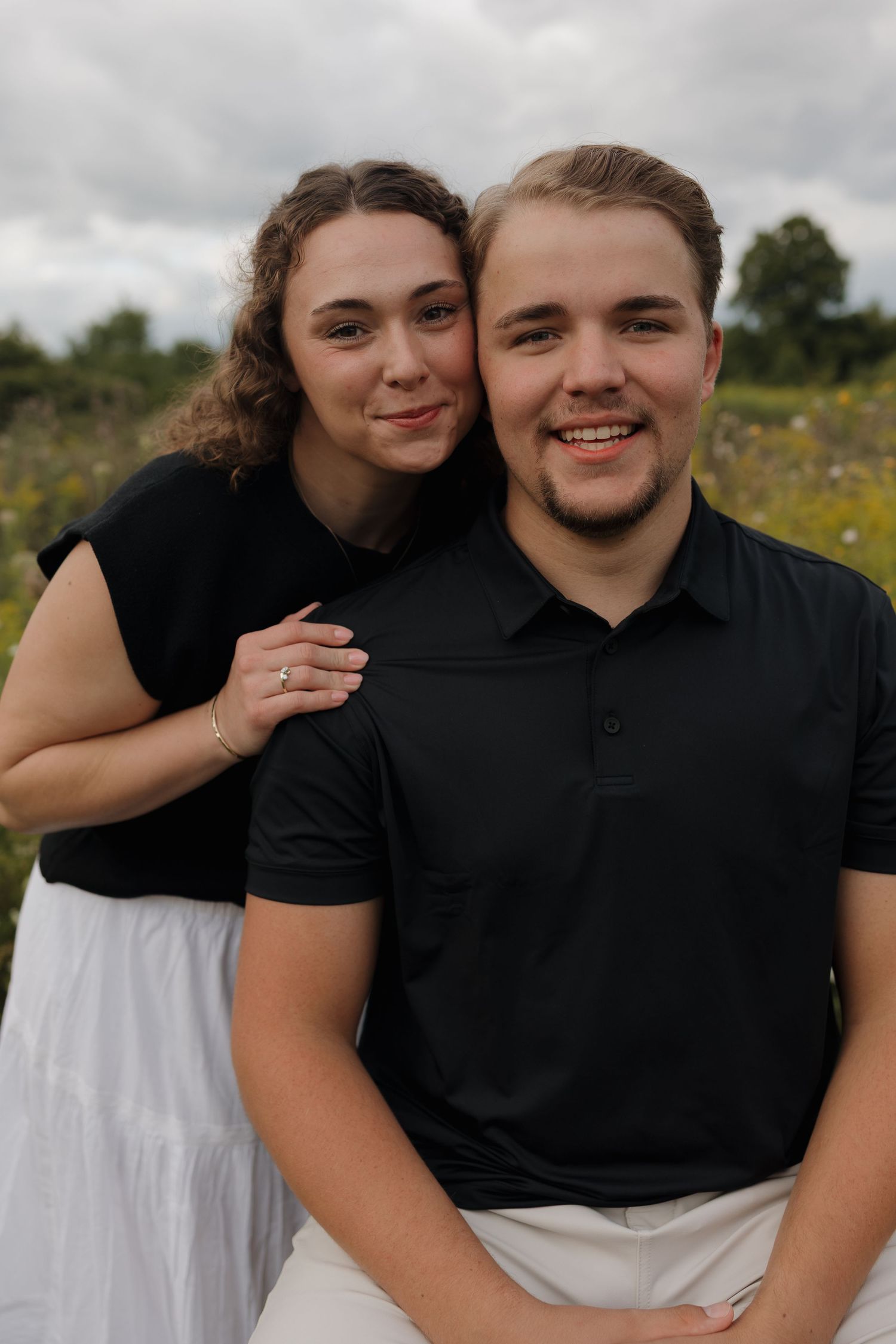 A smiling couple in matching black tops poses together in a flower field, the woman resting her head on the man's shoulder.