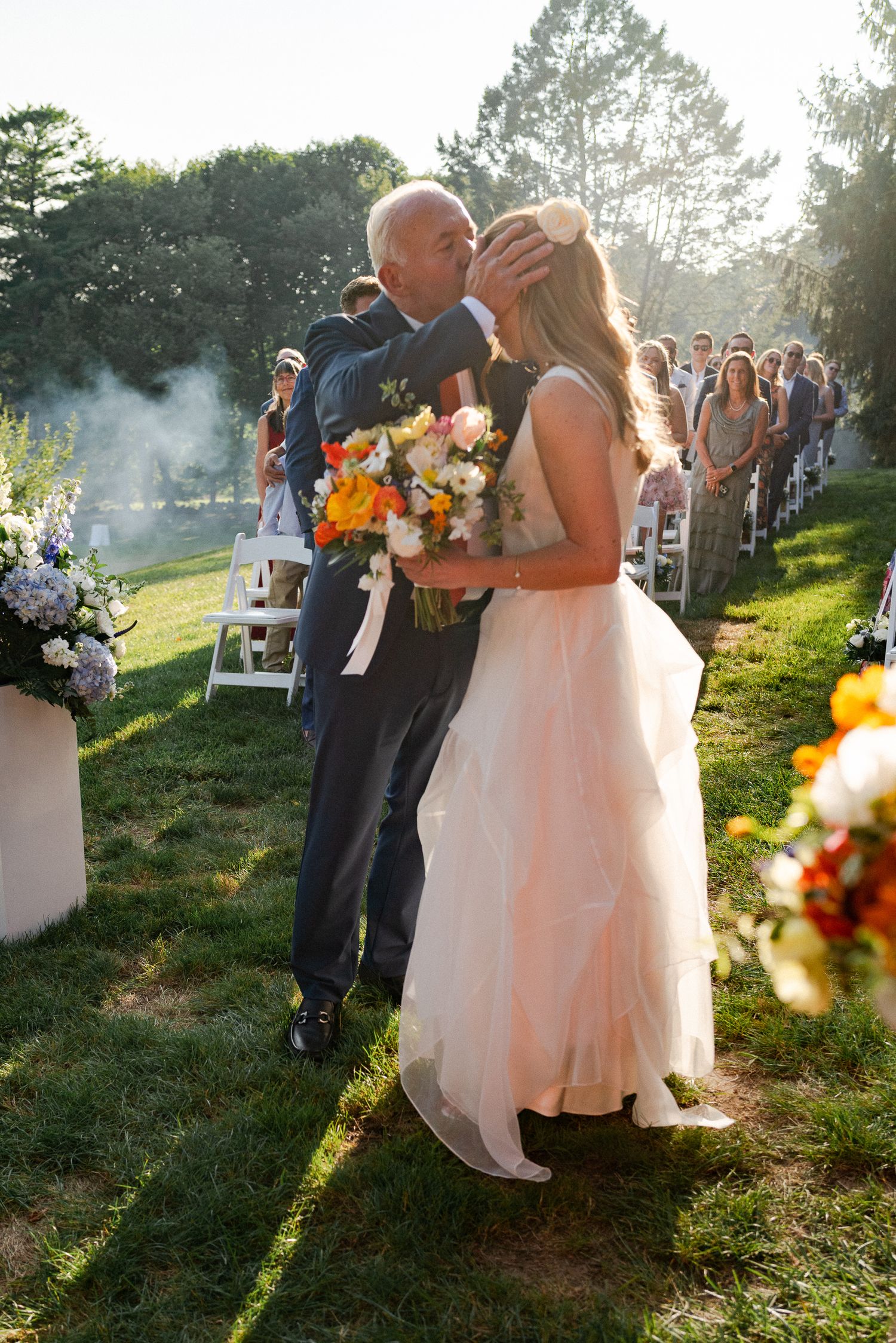 Father kissing bride on the forehead as she holds a vibrant bouquet during outdoor wedding ceremony.