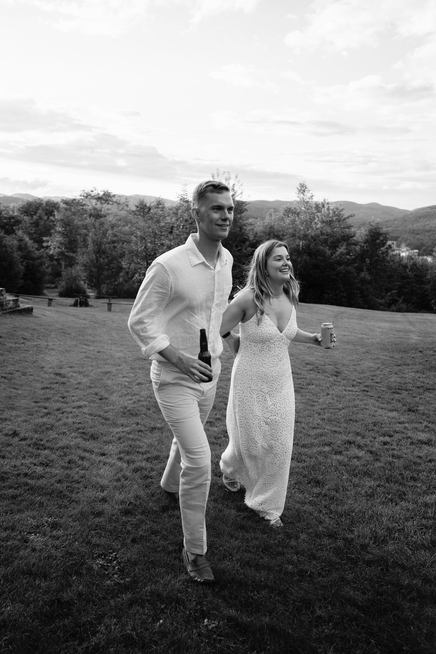 Black and white photo of a bride and groom walking together on a grassy field at dusk.