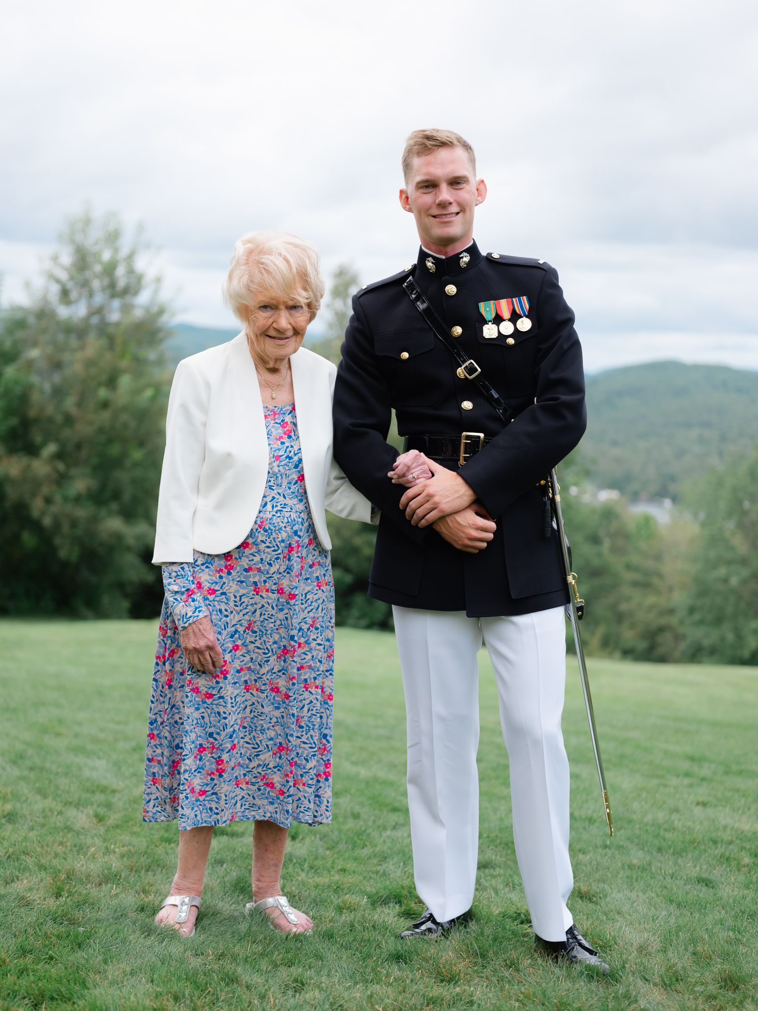 Marine in dress blues stands with elderly woman on green hillside with mountain views.