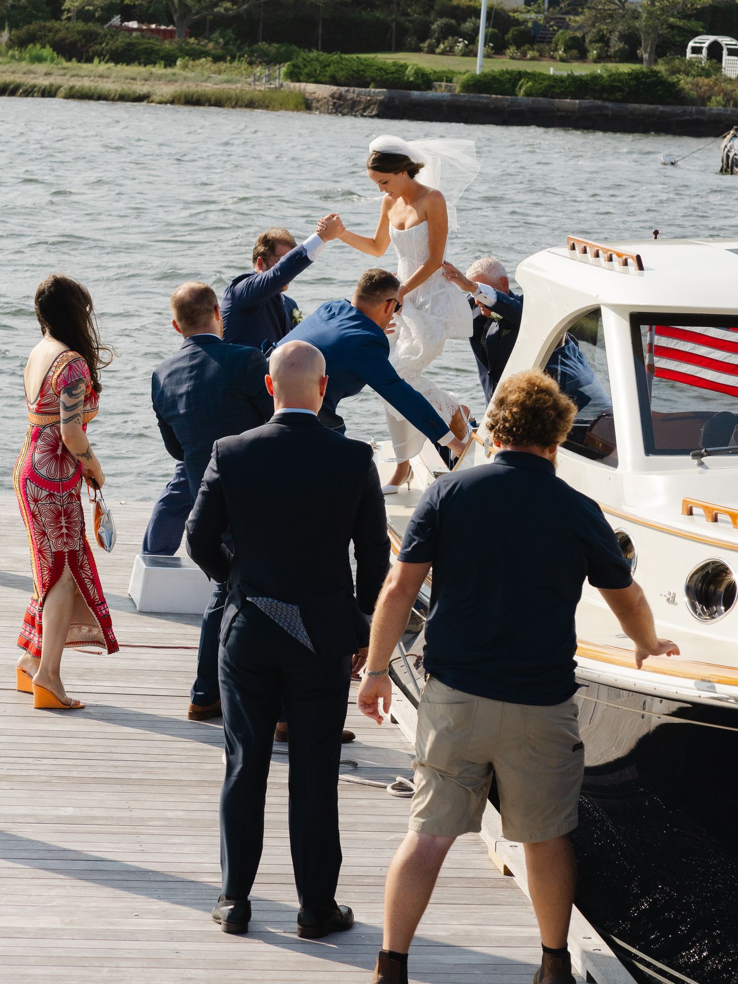 Wedding party helping bride board classic boat at dock after outdoor waterfront ceremony.