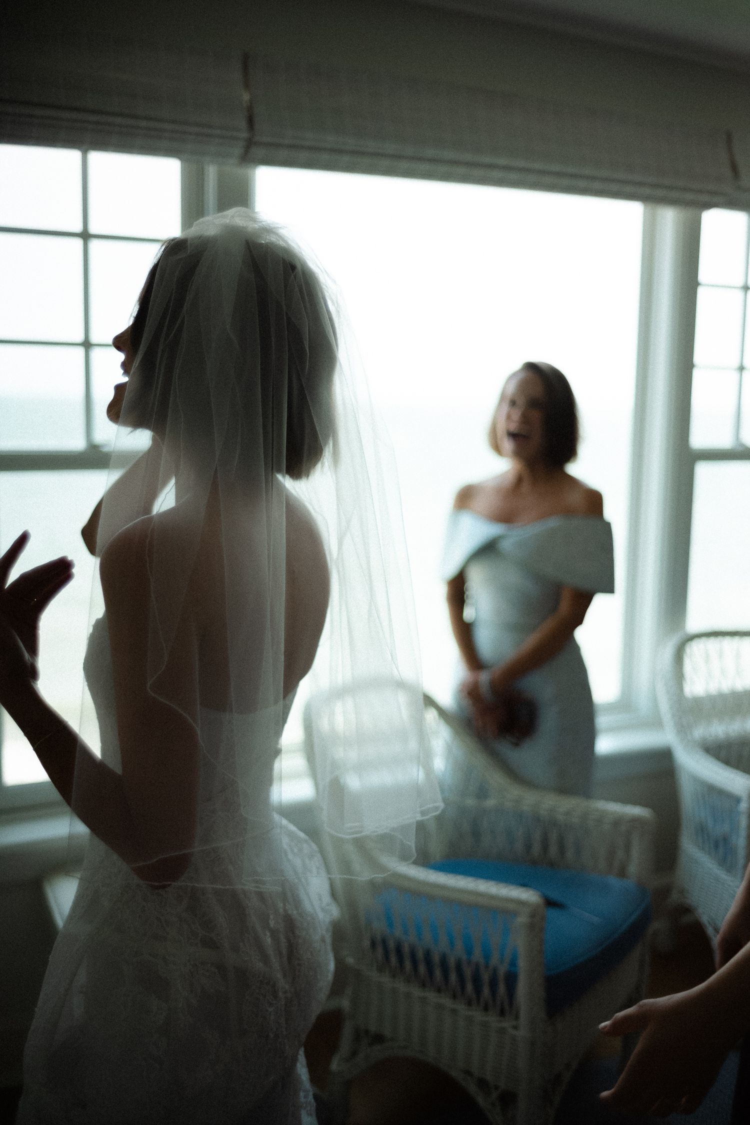 Bride in lace gown and veil shares emotional moment with woman in blue dress by window.