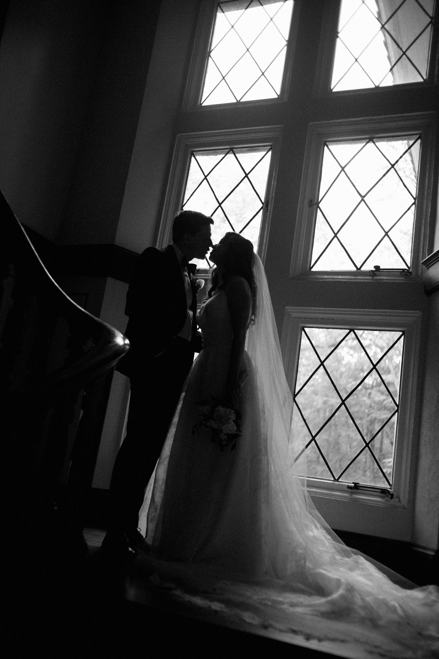 Silhouette of bride and groom kissing before large diamond-paned windows in a classic black and white wedding photo.