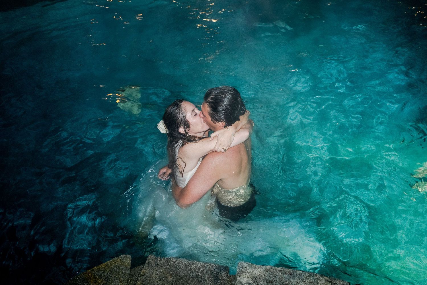 Bride and groom sharing romantic kiss while embracing in glowing turquoise pool at night.