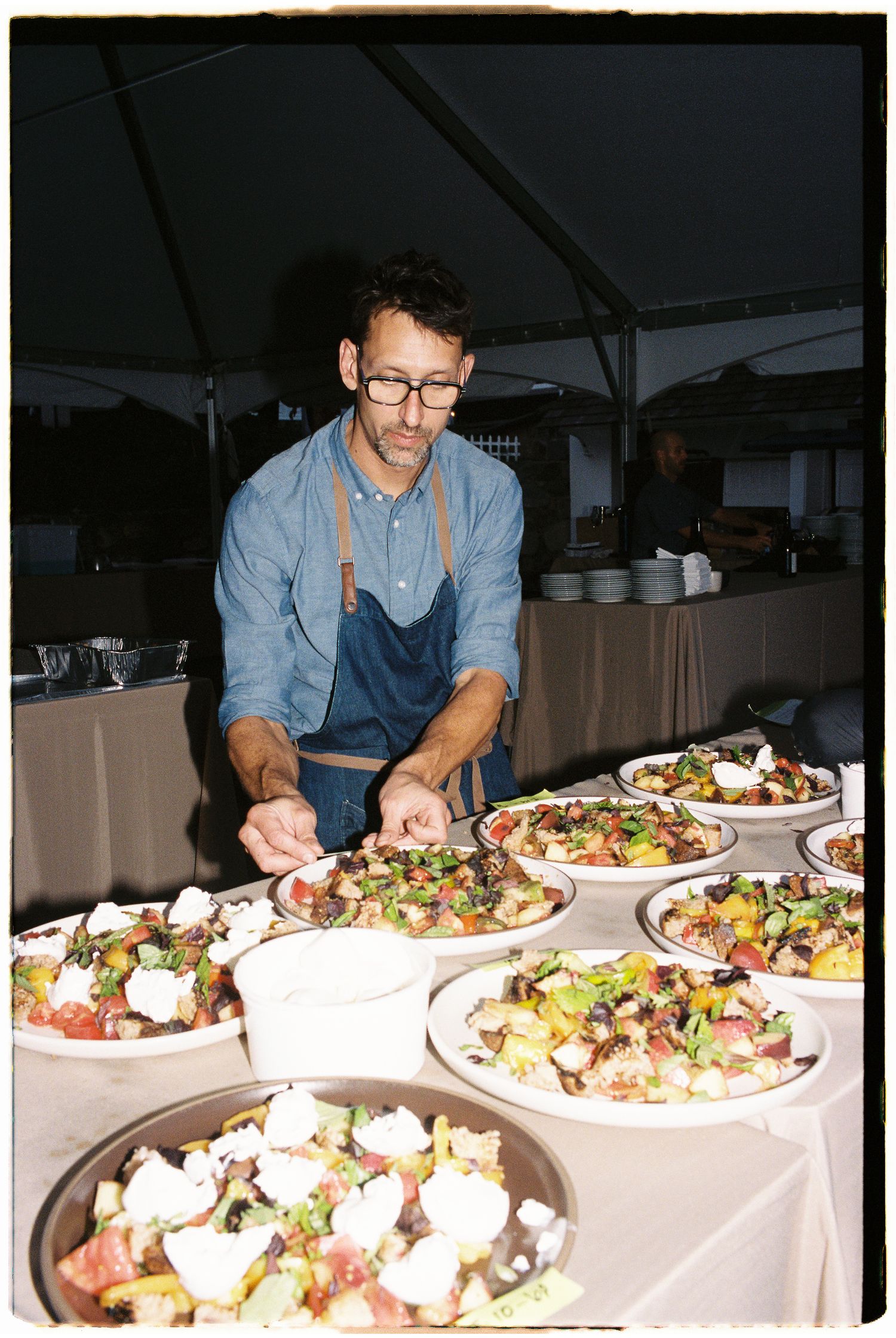 A chef in a blue apron and glasses plates colorful salad dishes at an outdoor catering event.
