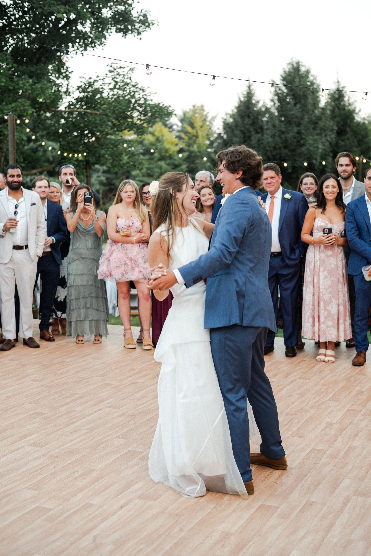 Bride and groom sharing their first dance on an outdoor wooden dance floor surrounded by wedding guests.