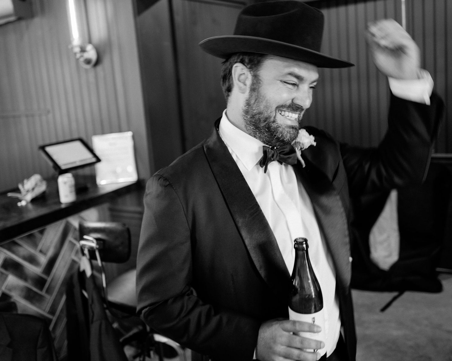 Groom in black tuxedo and wide-brim hat laughing while holding a beer bottle at wedding reception.
