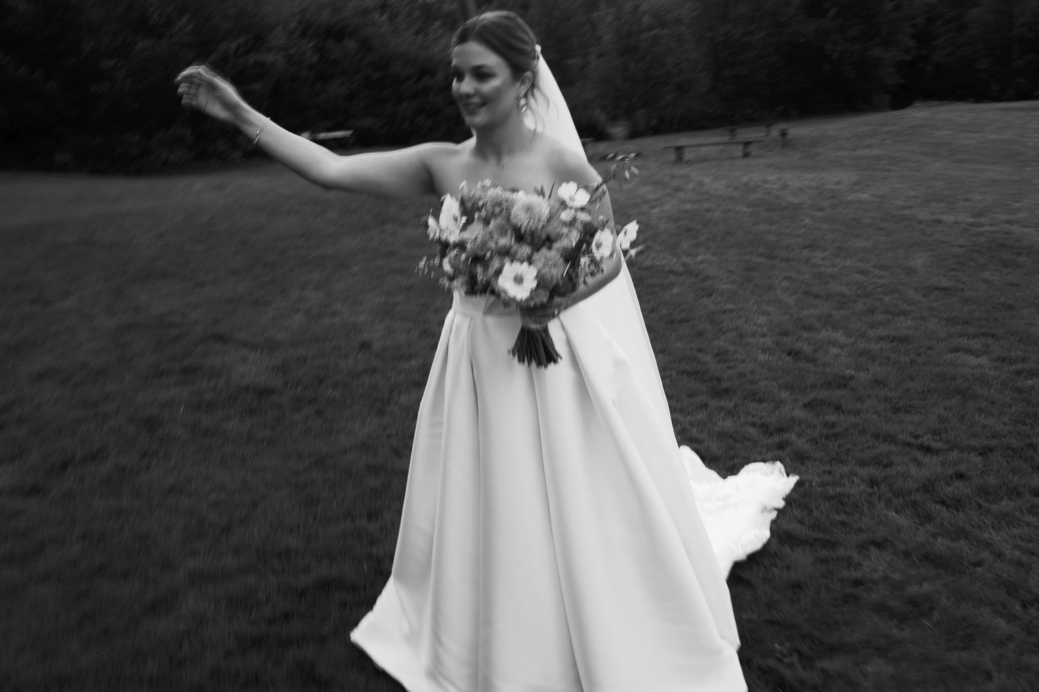 Black and white photo of bride in flowing white gown holding a bouquet in an open green field.