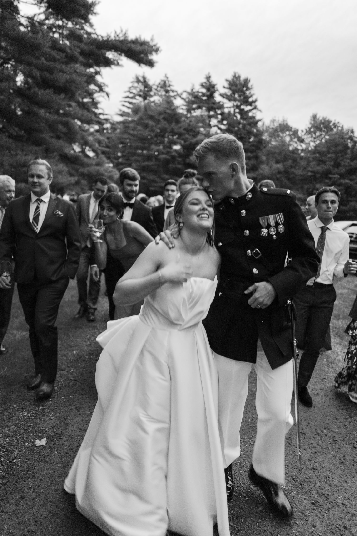 Bride in strapless white gown laughs joyfully with groom in Marine dress uniform surrounded by wedding guests.