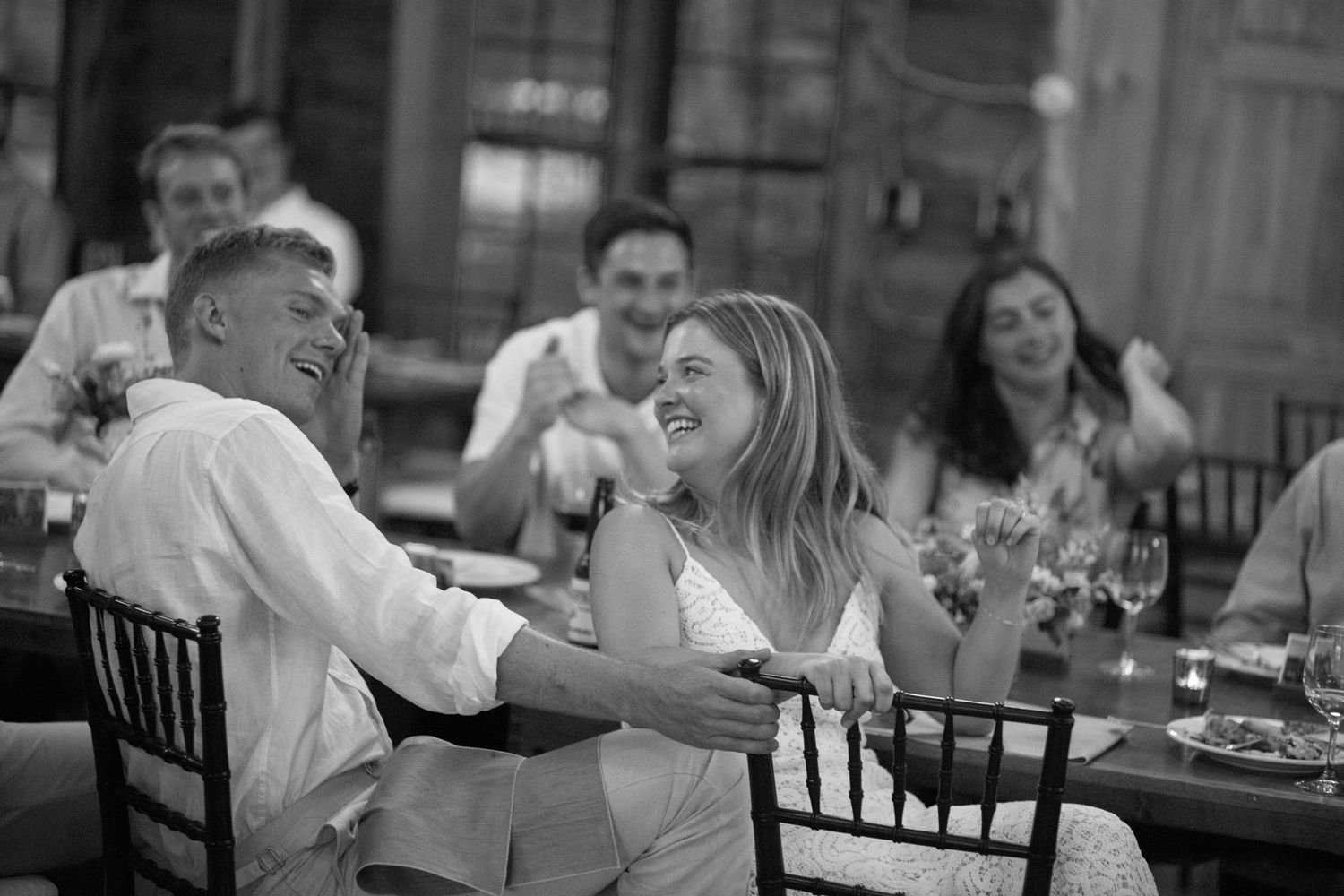 Black and white photo of a laughing bride and groom sharing a joyful moment at their reception table.