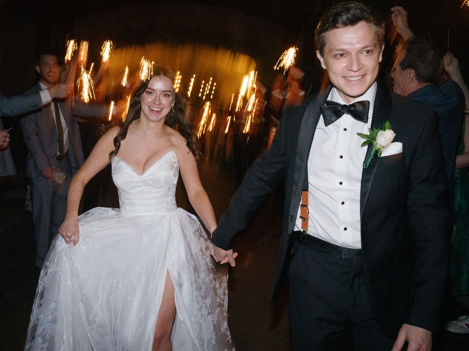 Smiling bride and groom hold hands walking through sparkler exit at wedding reception.