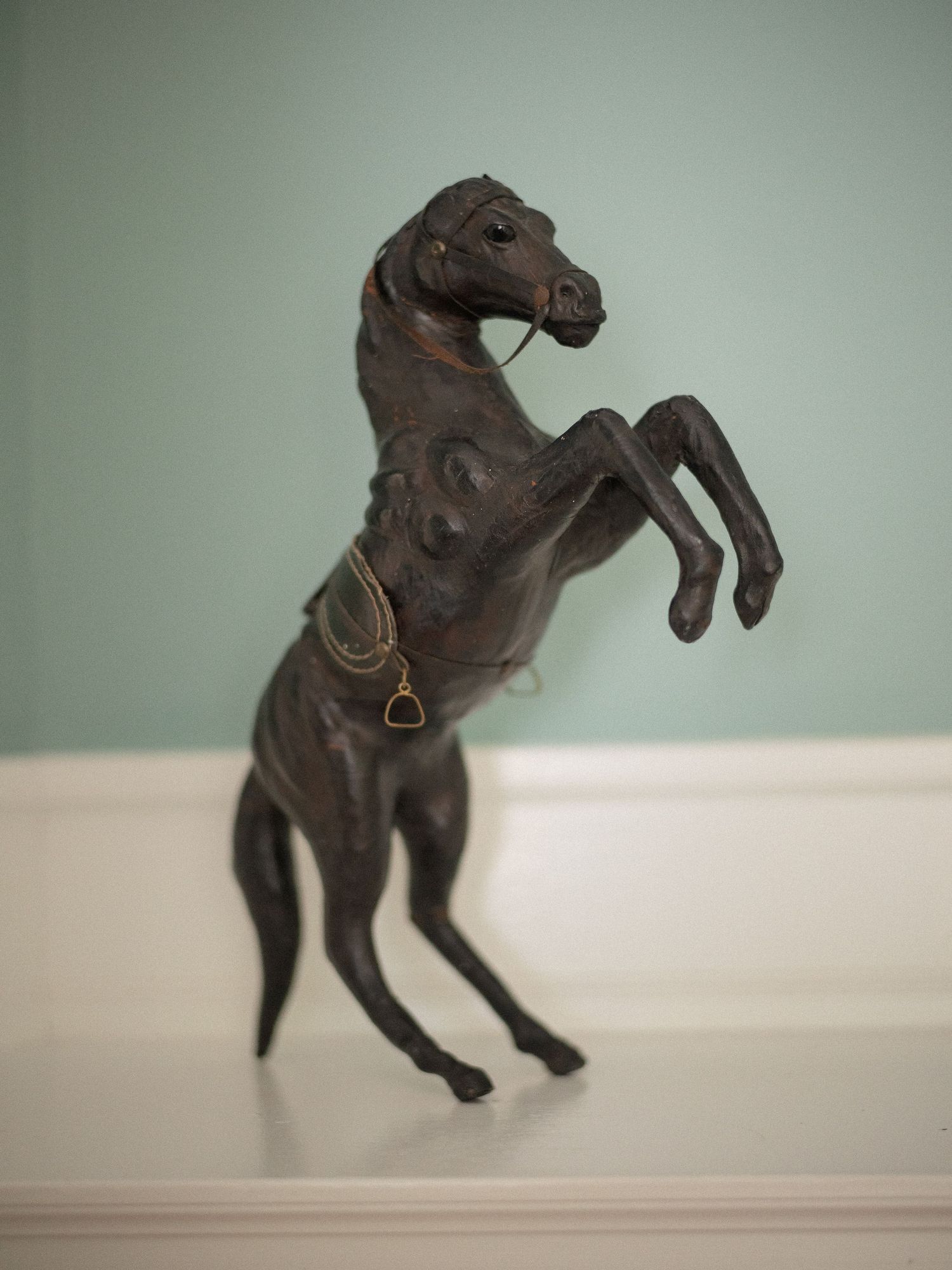 Dark bronze rearing horse figurine with gold stirrup detail displayed on white shelf against teal wall.